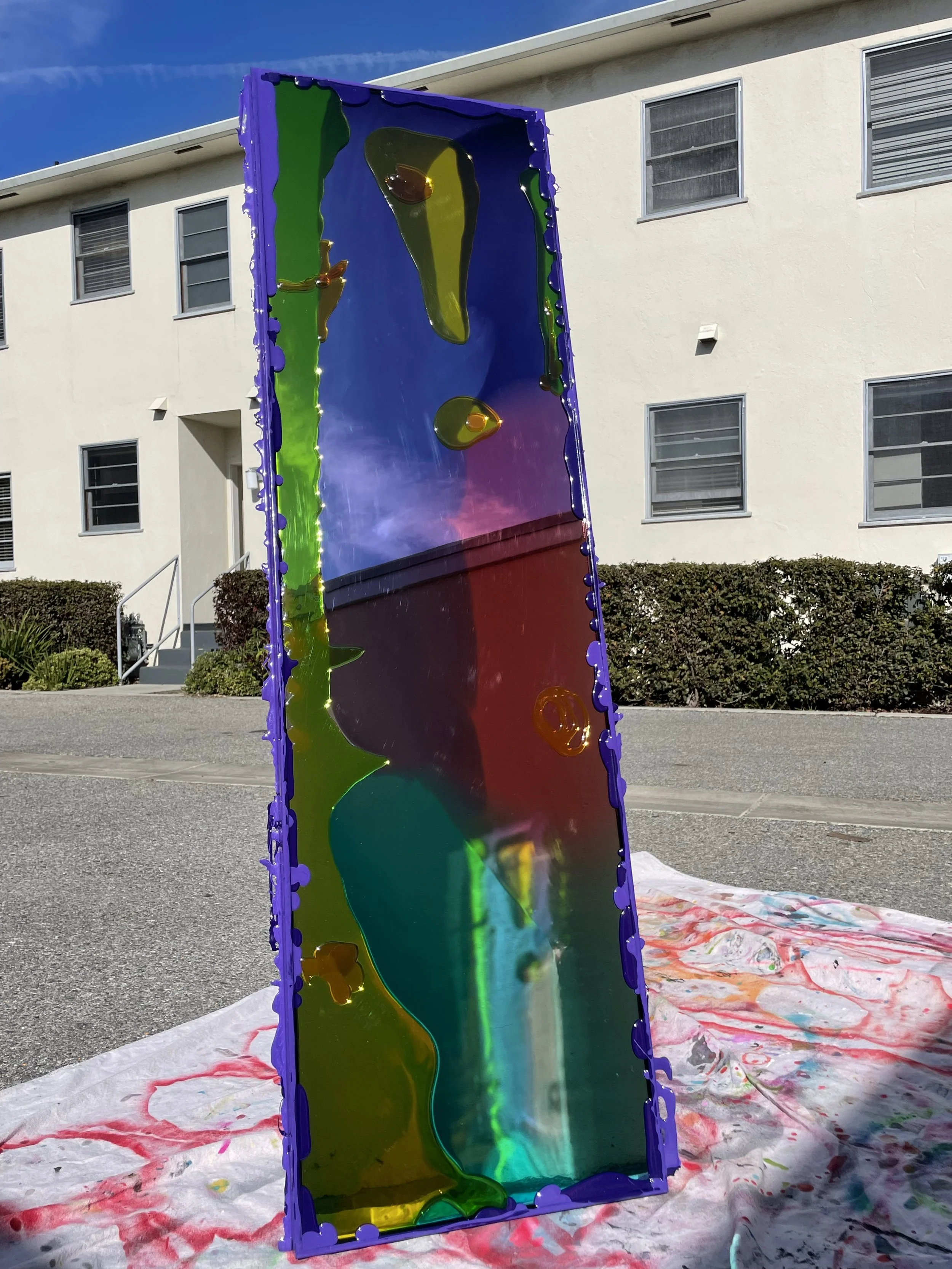 A tall, rectangular resin mirror with a colorful, abstract design and a purple border, placed outdoors on a sunny day with a residential building in the background.