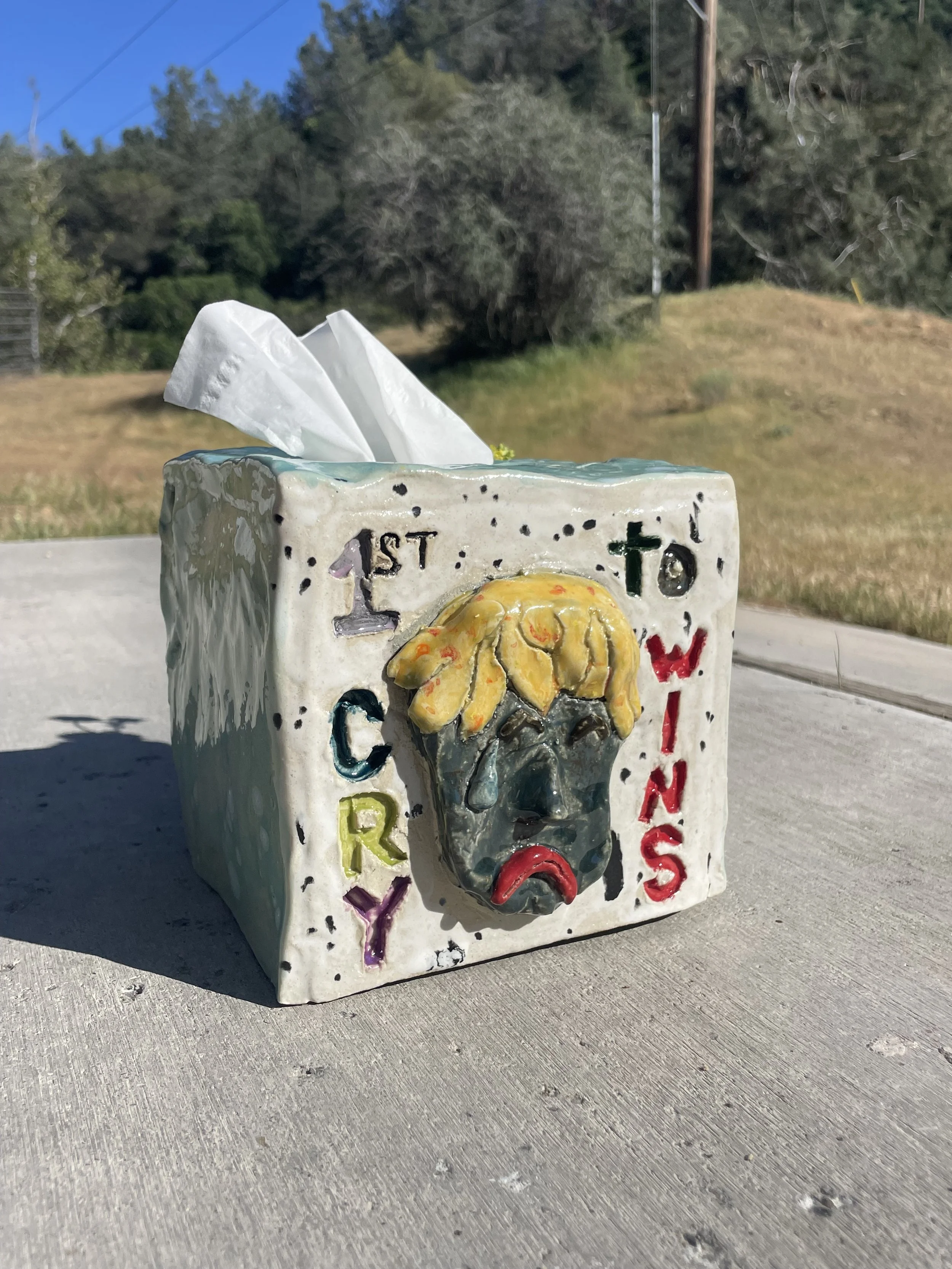 Decorative tissue box with the words 'First to Cry' and a sad face with tears, outdoors on a sidewalk.