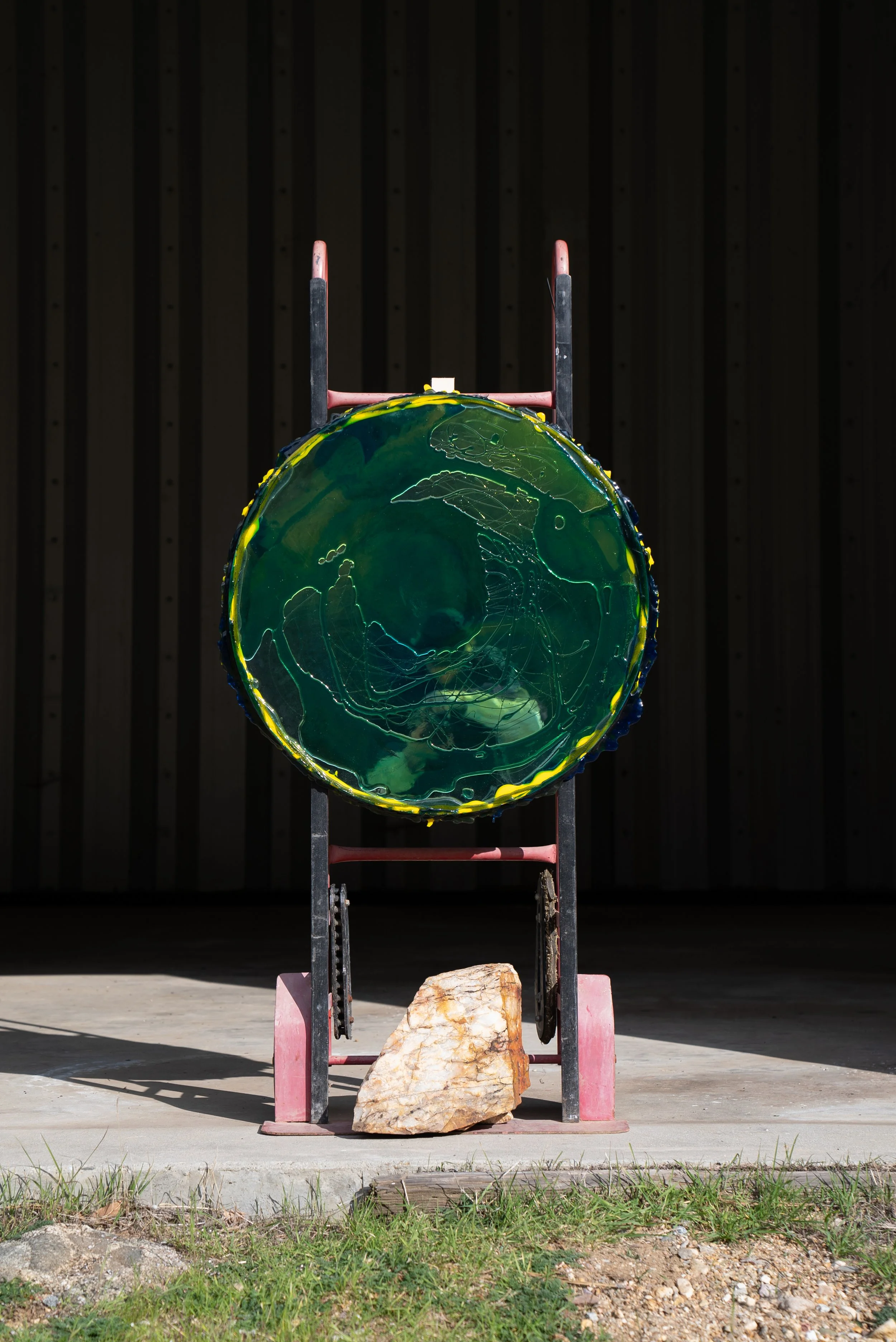 A circular stained glass-like resin mirror with green and yellow colors, mounted on a rack, with a large rock at its base, outdoors on a concrete surface with grass and a wooden wall background.