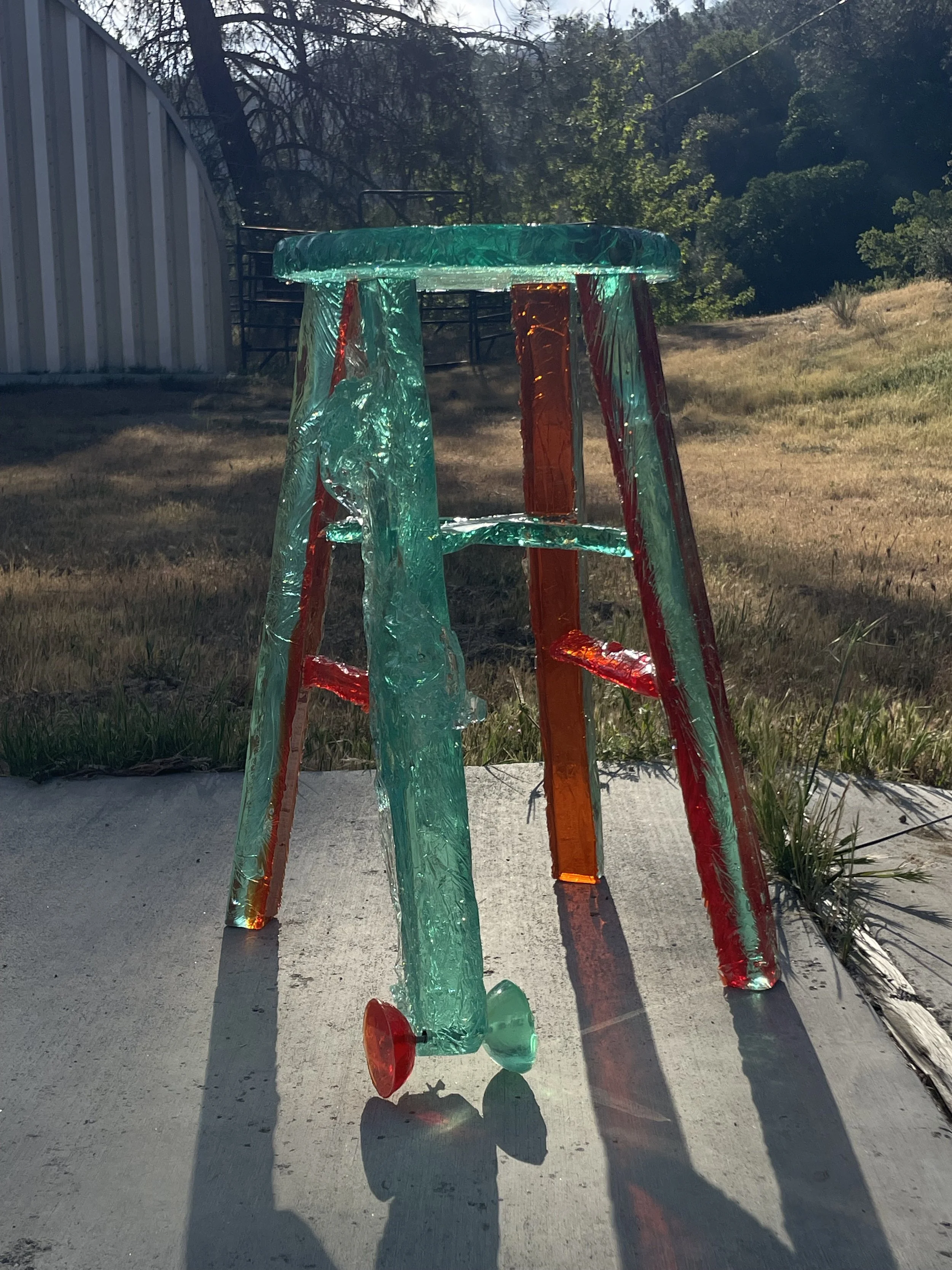 Glass art sculpture resembling a small dining table with four legs, made of colorful and jagged glass pieces, placed outdoors on a concrete surface with trees and grass in the background.
