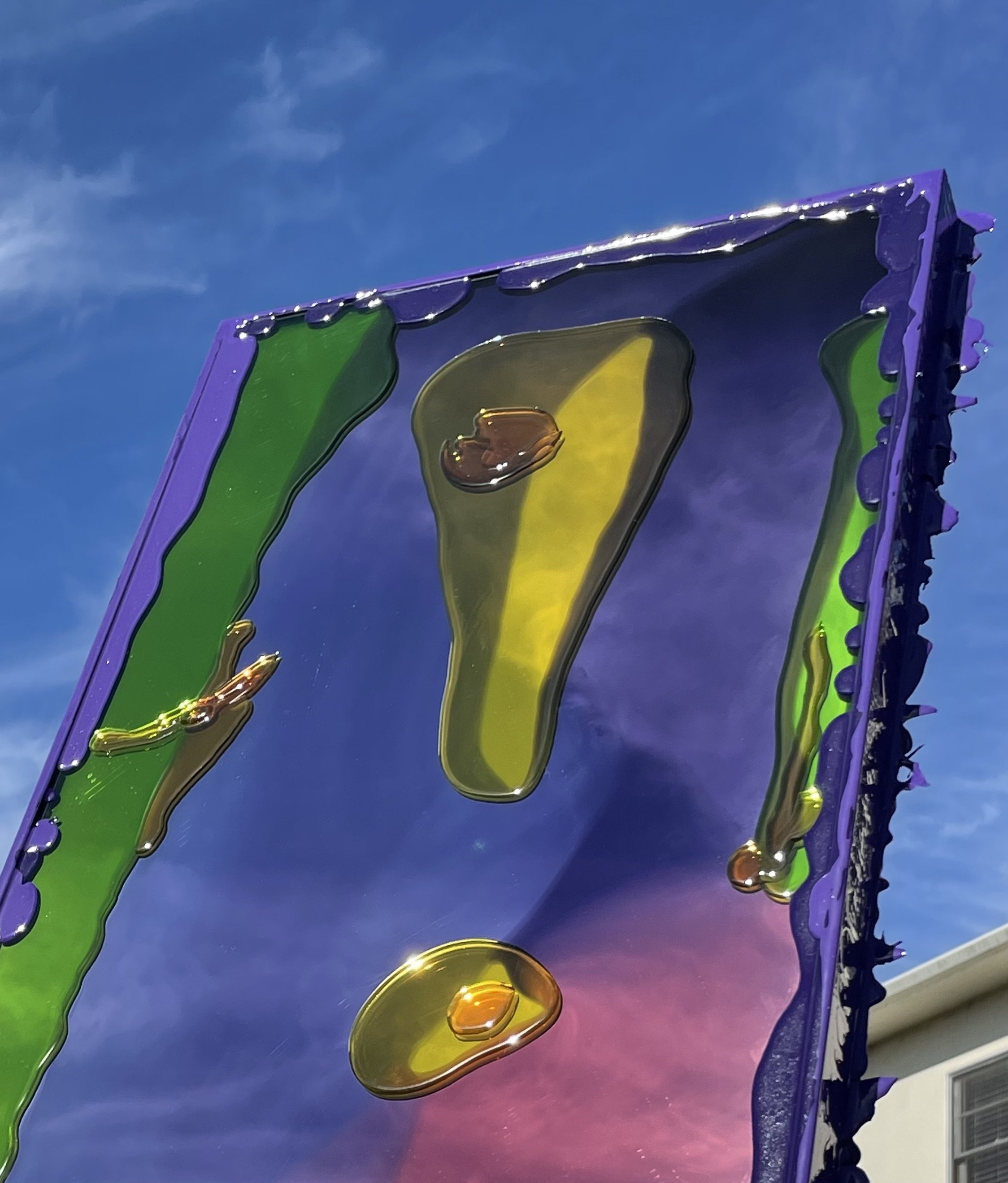 Colorful resin mirror with yellow, green, and purple sections, with droplets of resin on its surface, against a blue sky background.