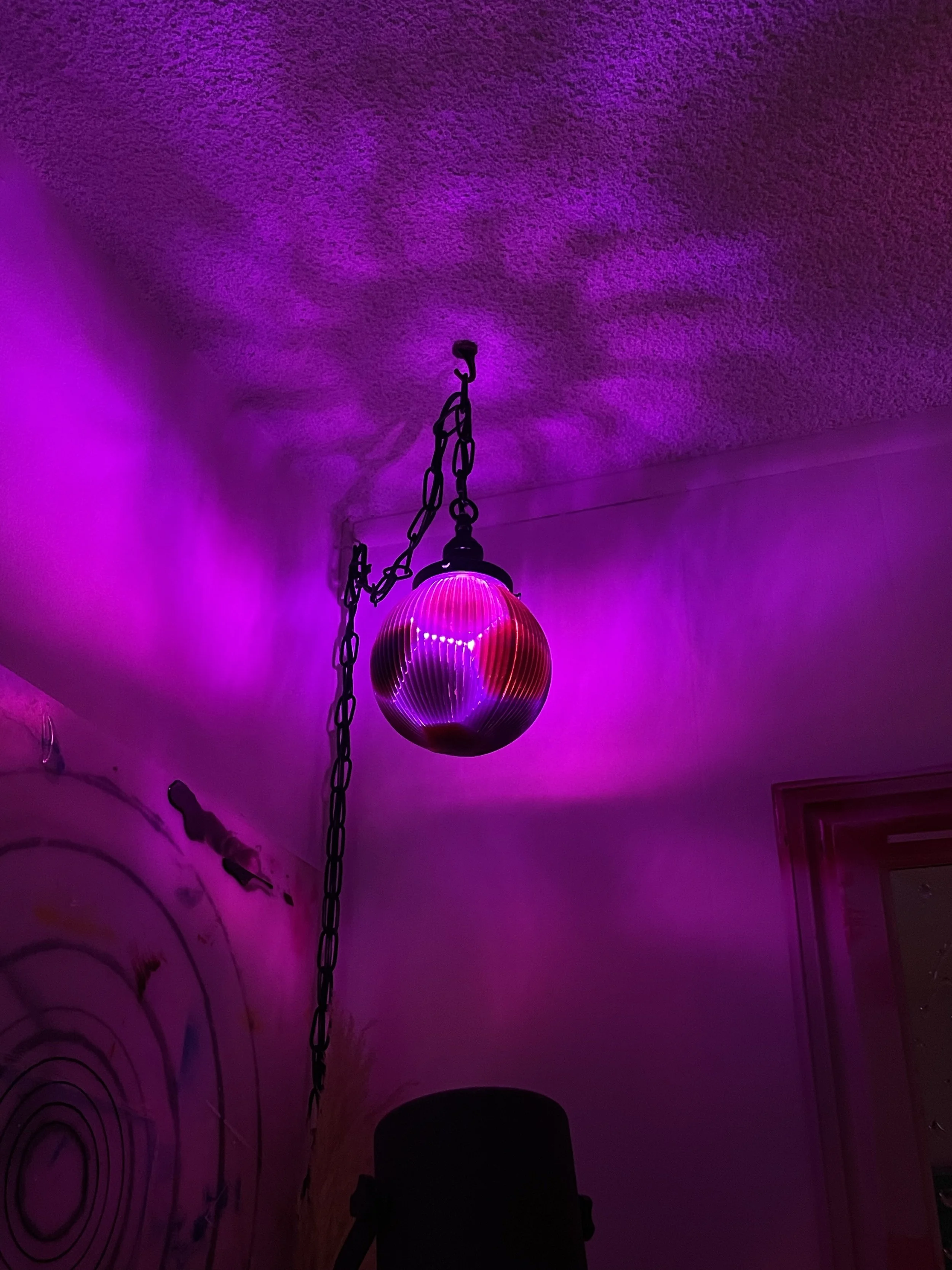A hanging resin lamp with a pinkish-purple light creates colorful reflections on a textured ceiling in a dimly lit room.