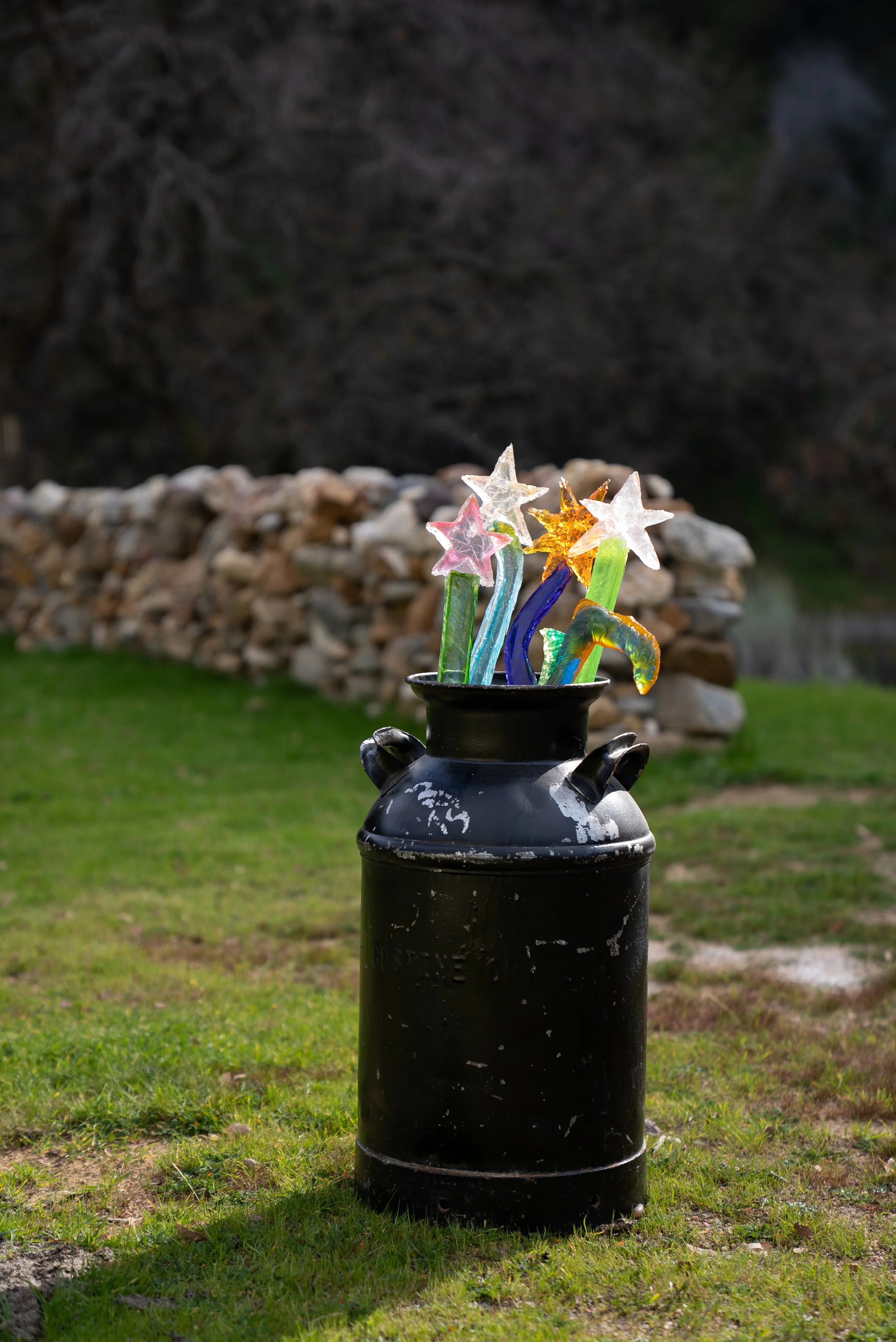 Colorful resin star-shaped magic wands in a black metal pot outdoors with a grassy area and a stone wall in the background.