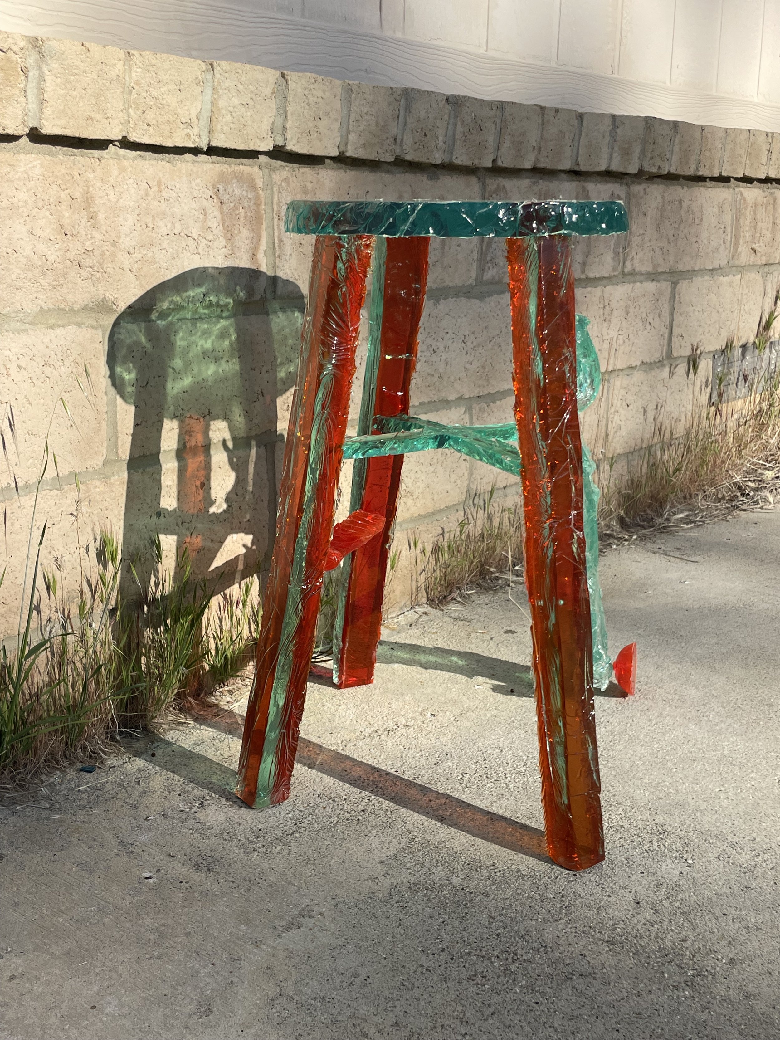 A colorful glass sculpture resembling a small table or stool, with a round top and four legs, leaning against a brick and concrete wall, casting shadows on the ground.