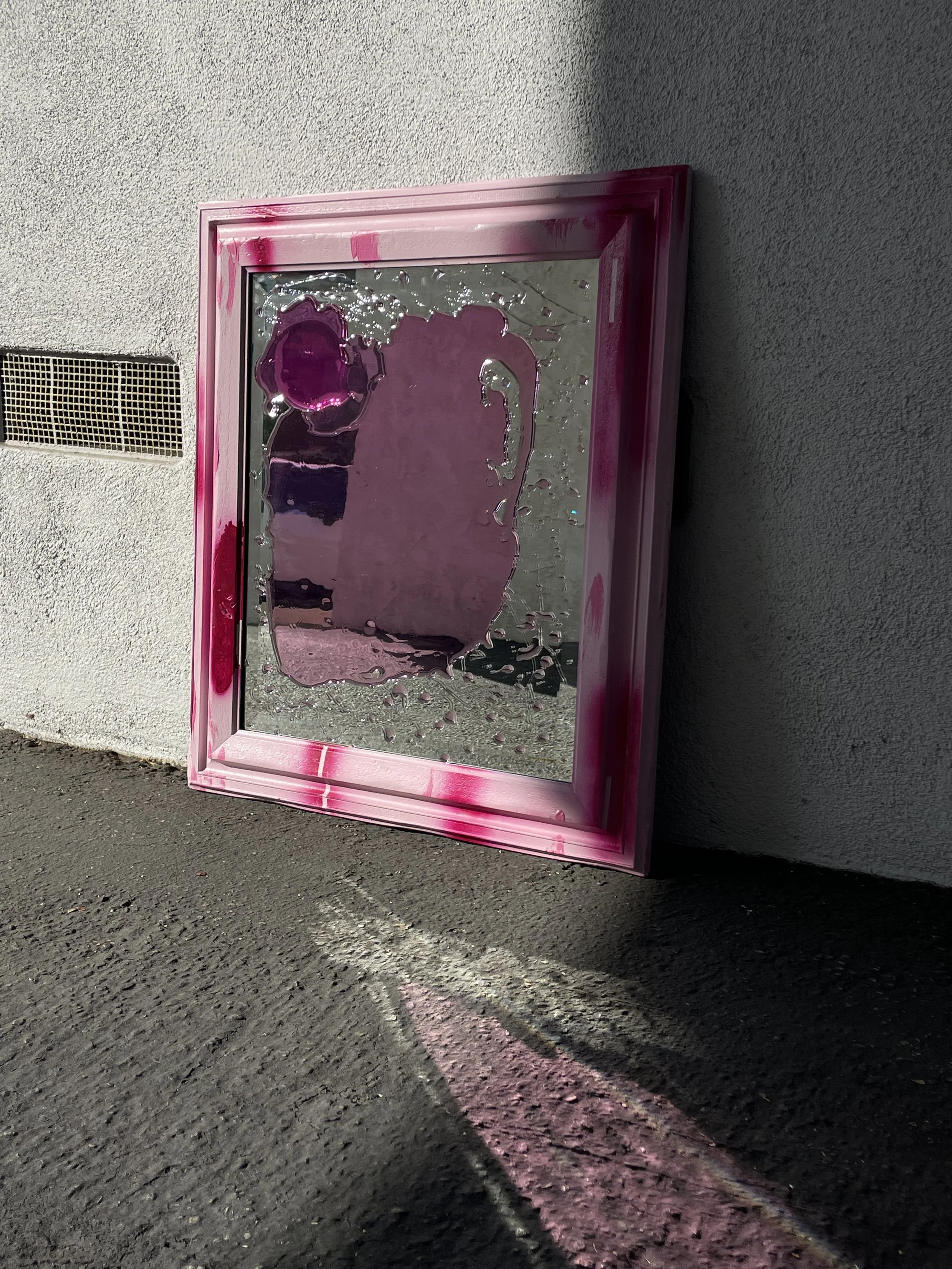 A pink-framed resin mirror leaning against a gray wall outdoors, casting pinkish shadows on the ground.