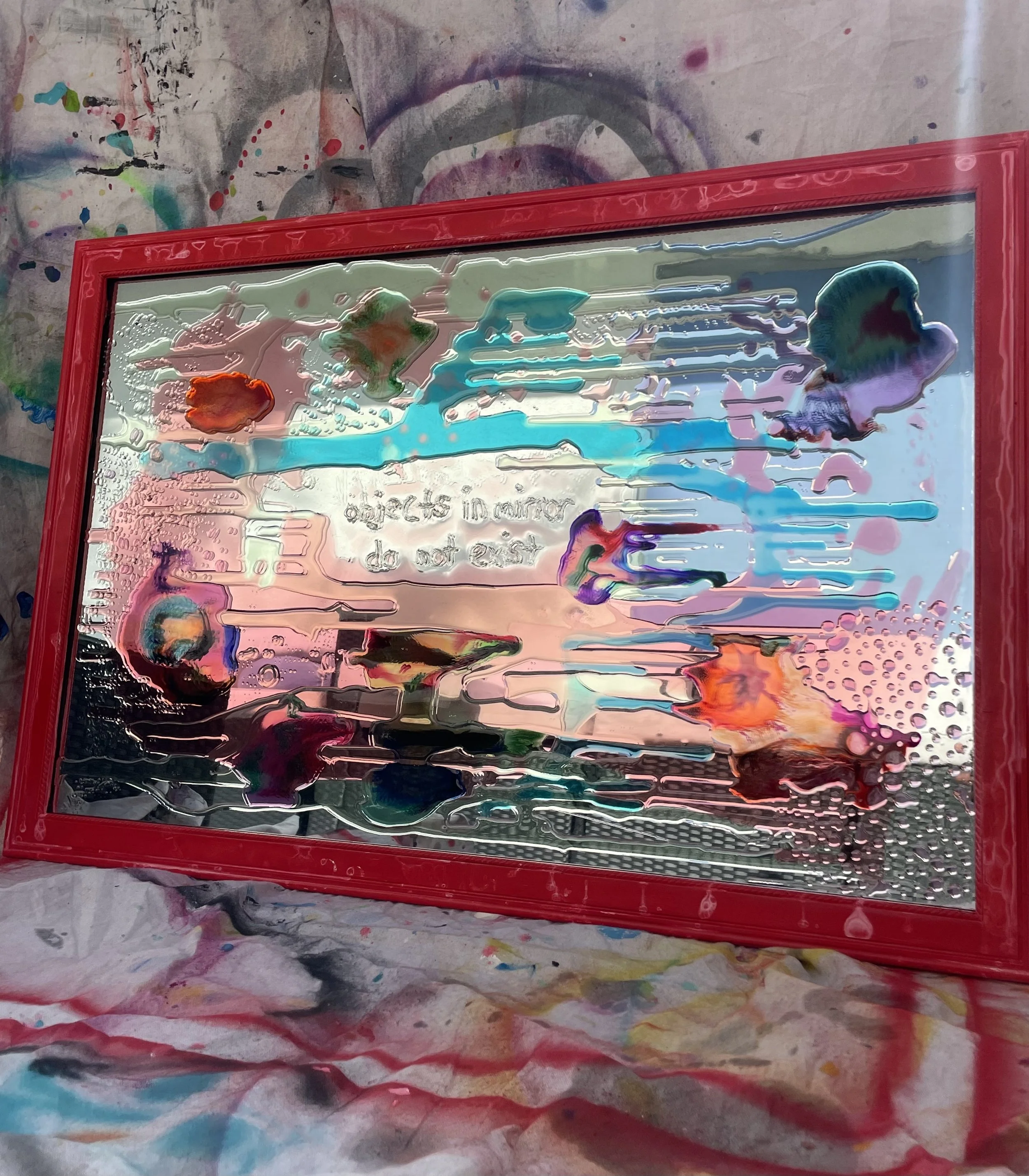 Resin representing paint splatters on a rectangular glass mirror with a red frame, with some writing visible through the glass, and is placed on a colorful, paint-splattered cloth background.