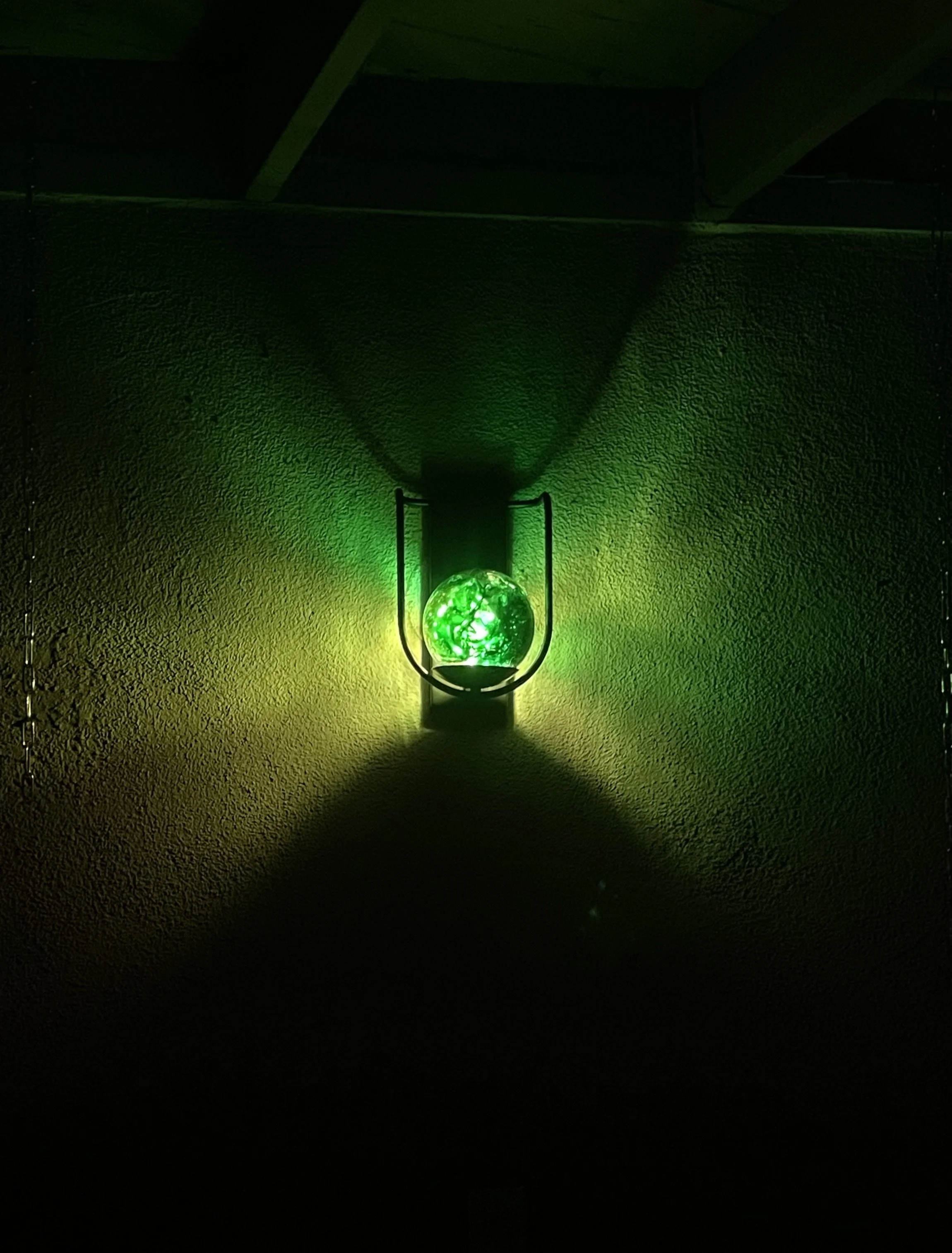 A glowing green resin orb with swirling patterns, mounted on a black stand against a textured wall, casting green and yellow light shadows.