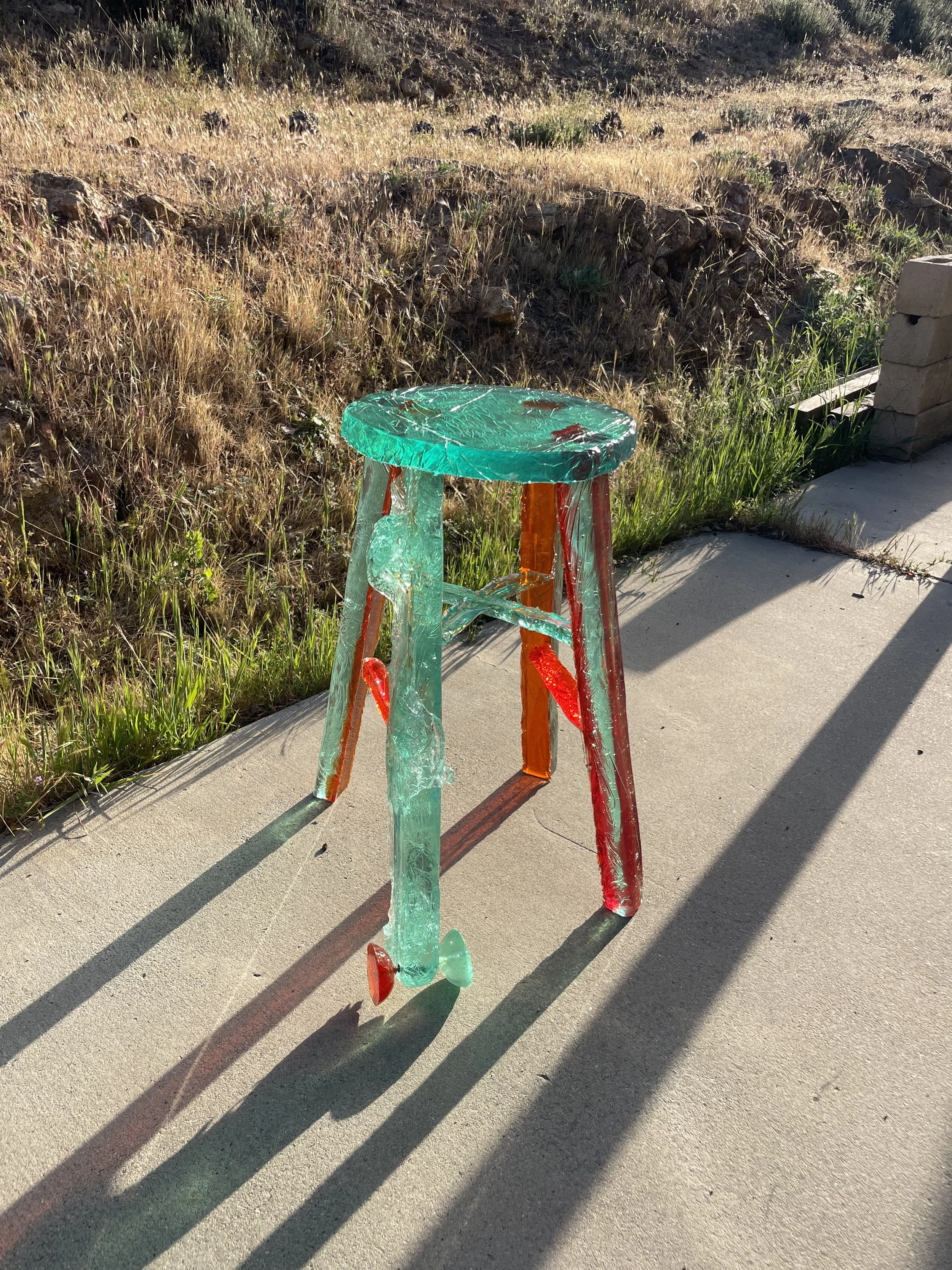 A chair made of shattered glass sits on a sidewalk with a grassy and rocky hillside in the background, casting a long shadow.