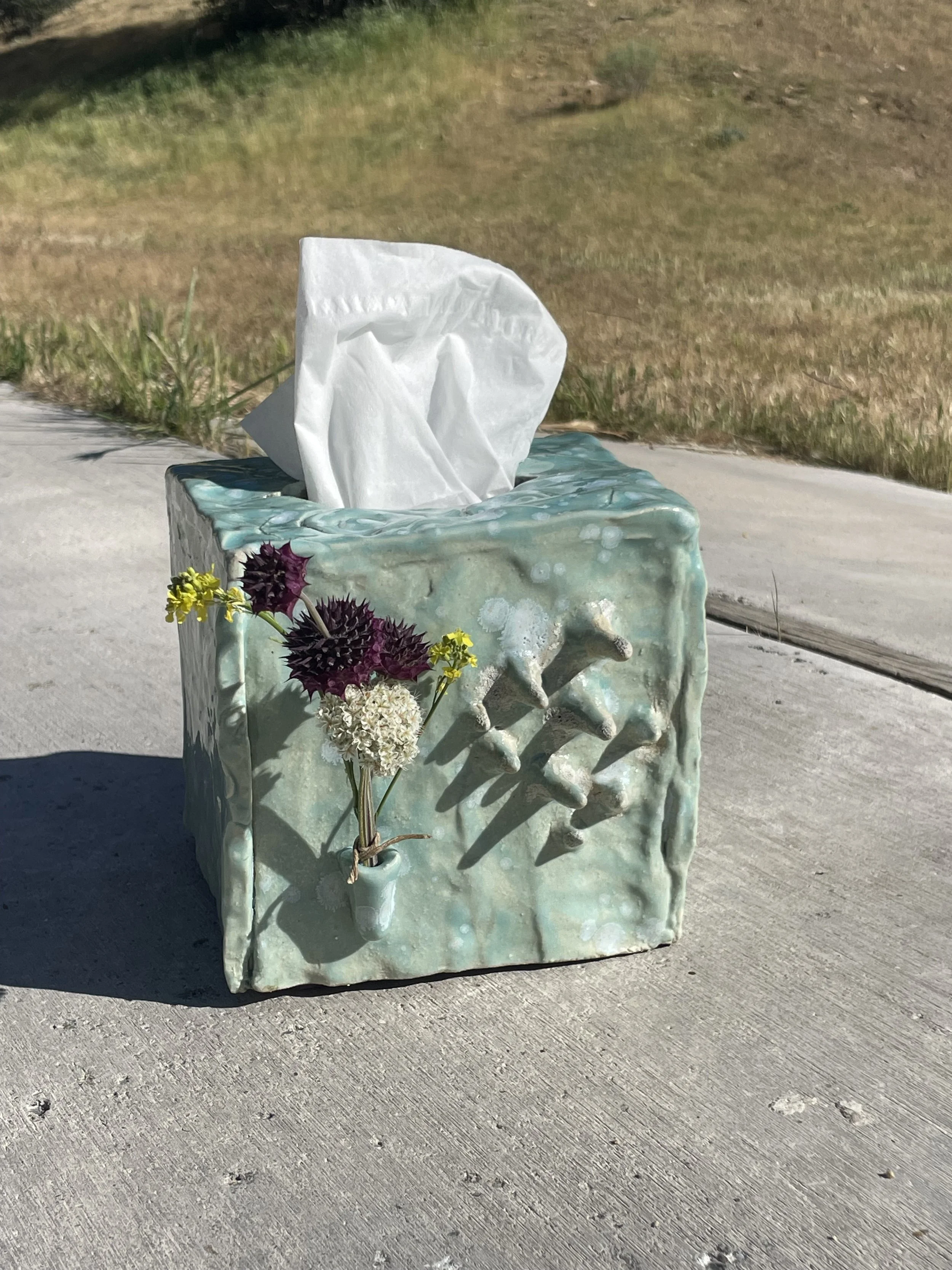 Ceramic tissue box holder with a small bouquet of dried flowers attached, placed on a concrete surface outdoors.