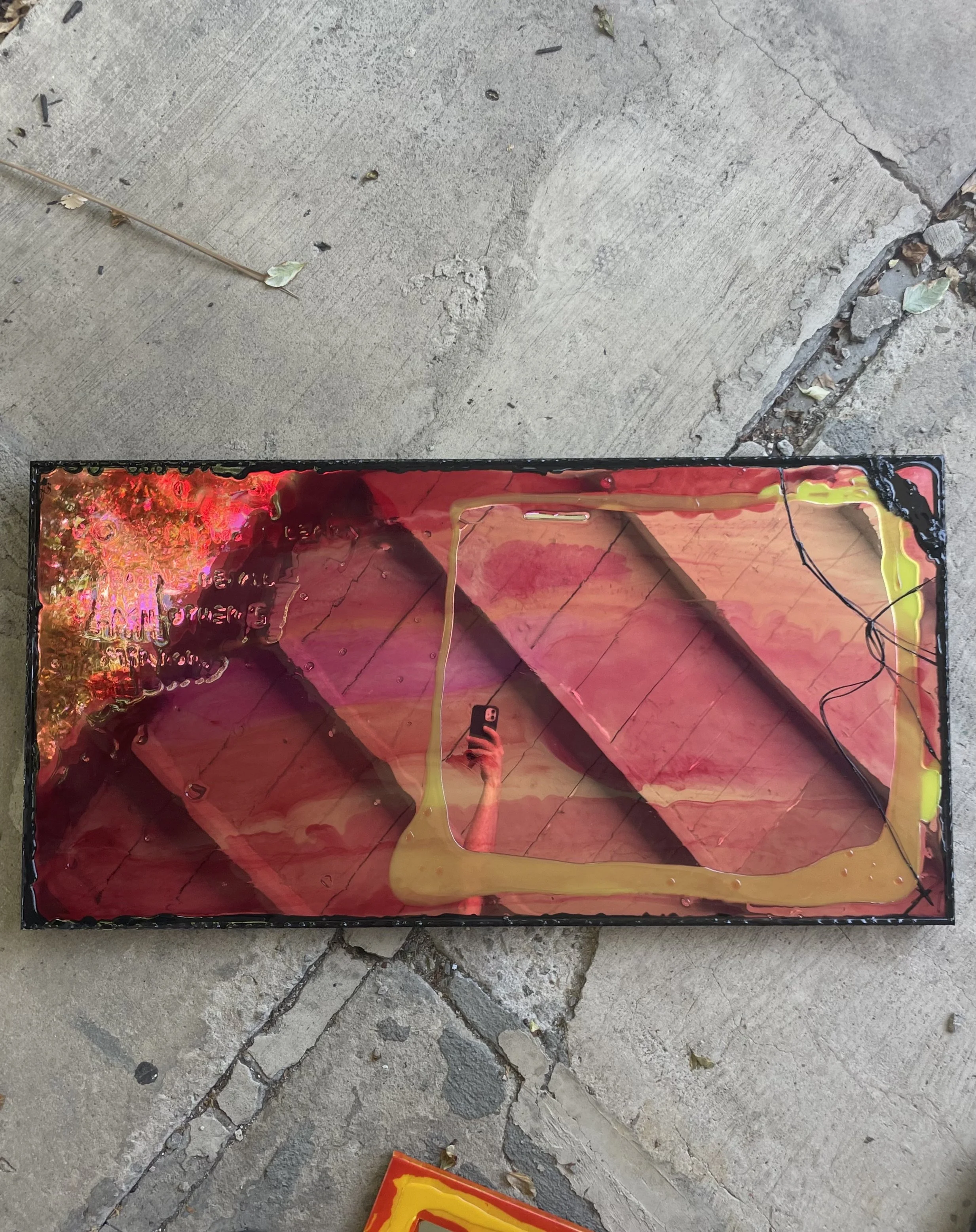 A rectangular resin mirror with a colorful, abstract design, placed on a concrete ground. The design features red, pink, orange, and yellow hues with dark lines, and the reflection of the person taking the photo can be seen in the glass.