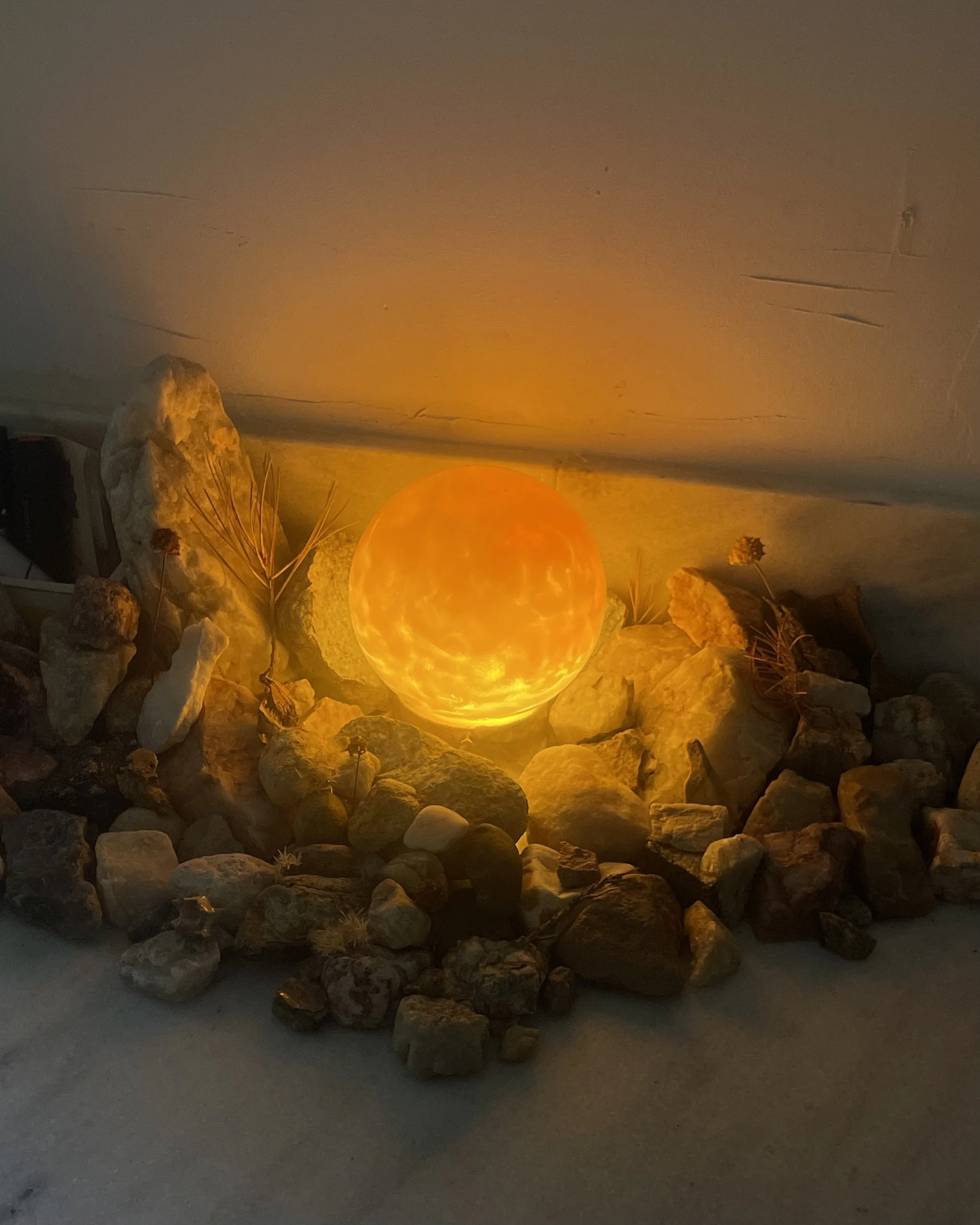 Illuminated orange spherical resin lamp surrounded by rocks and small dried plants.
