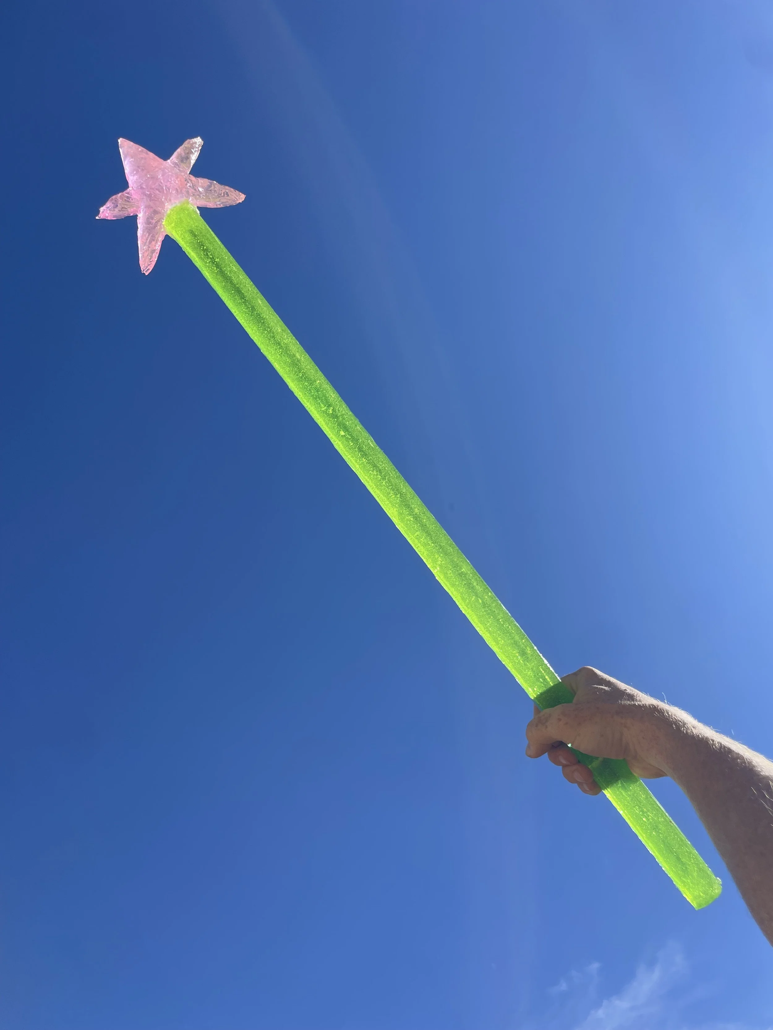Hand holding a green and pink resin fairy wand against a blue sky.