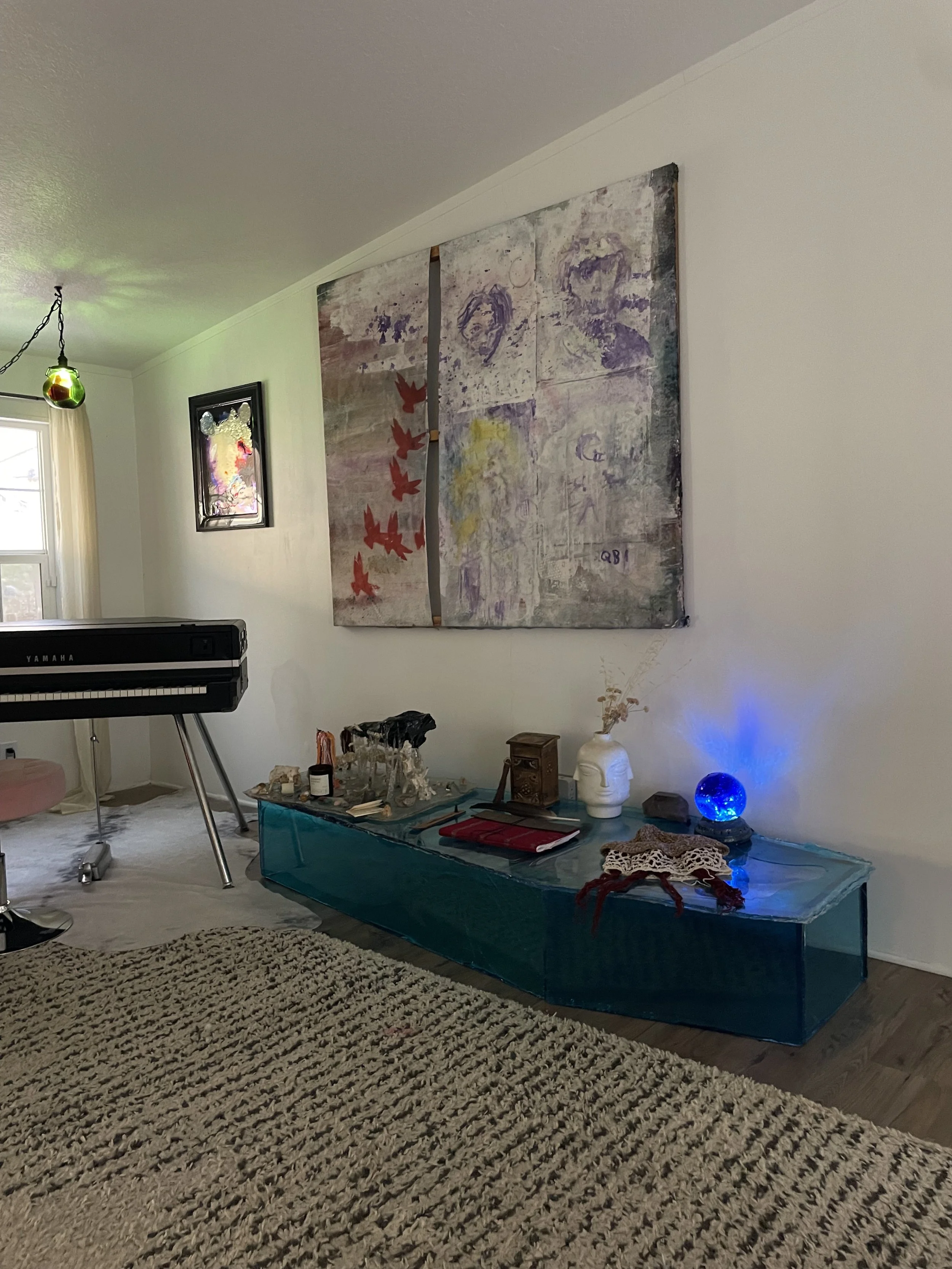 Living room with abstract wall art, Yamaha piano, decorative objects on a blue glass-like resin coffin coffee table, and a patterned rug.
