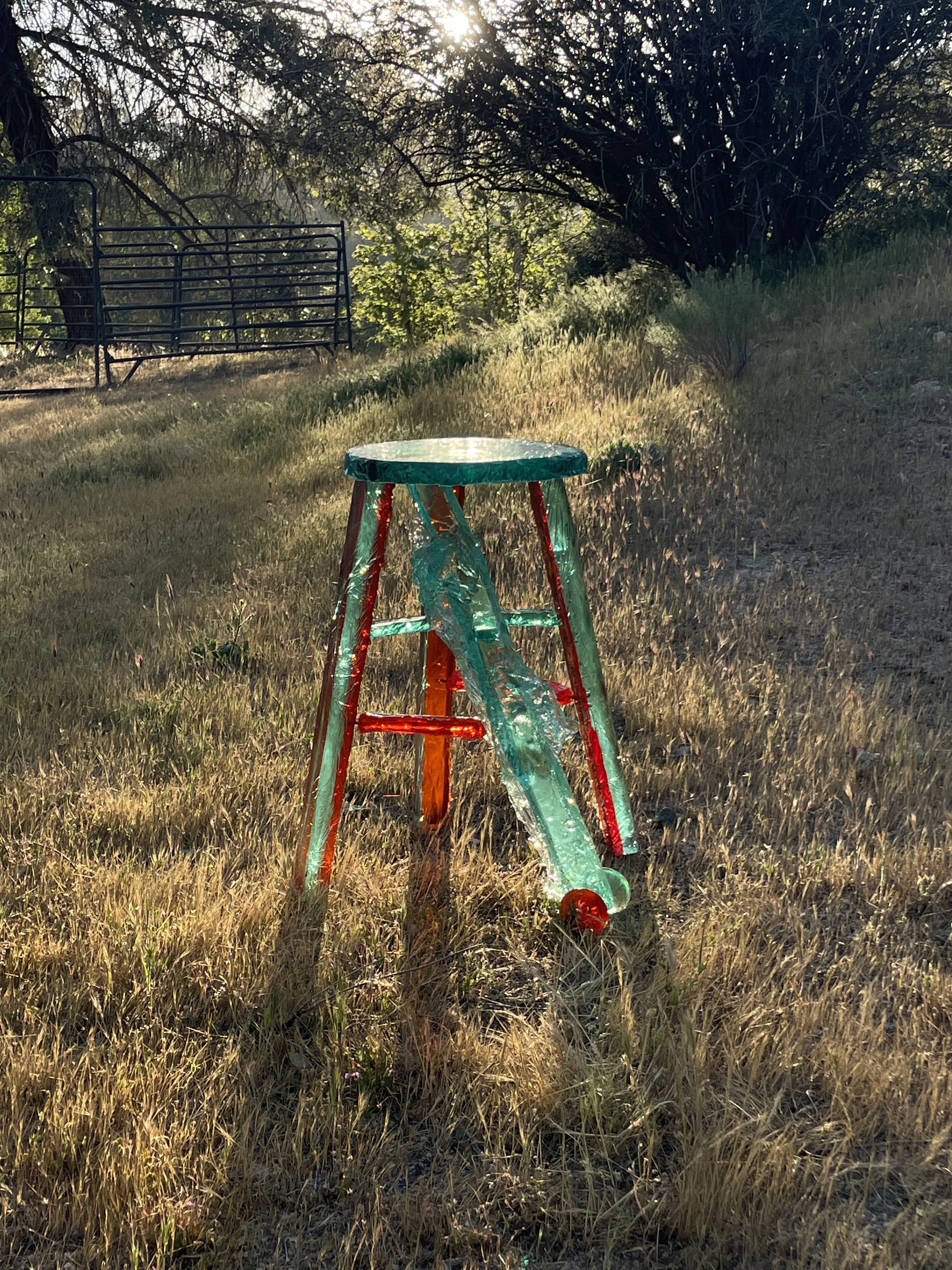 Colorful resin stool on a grassy field with trees and a metal fence in the background, sunlight shining through the trees.
