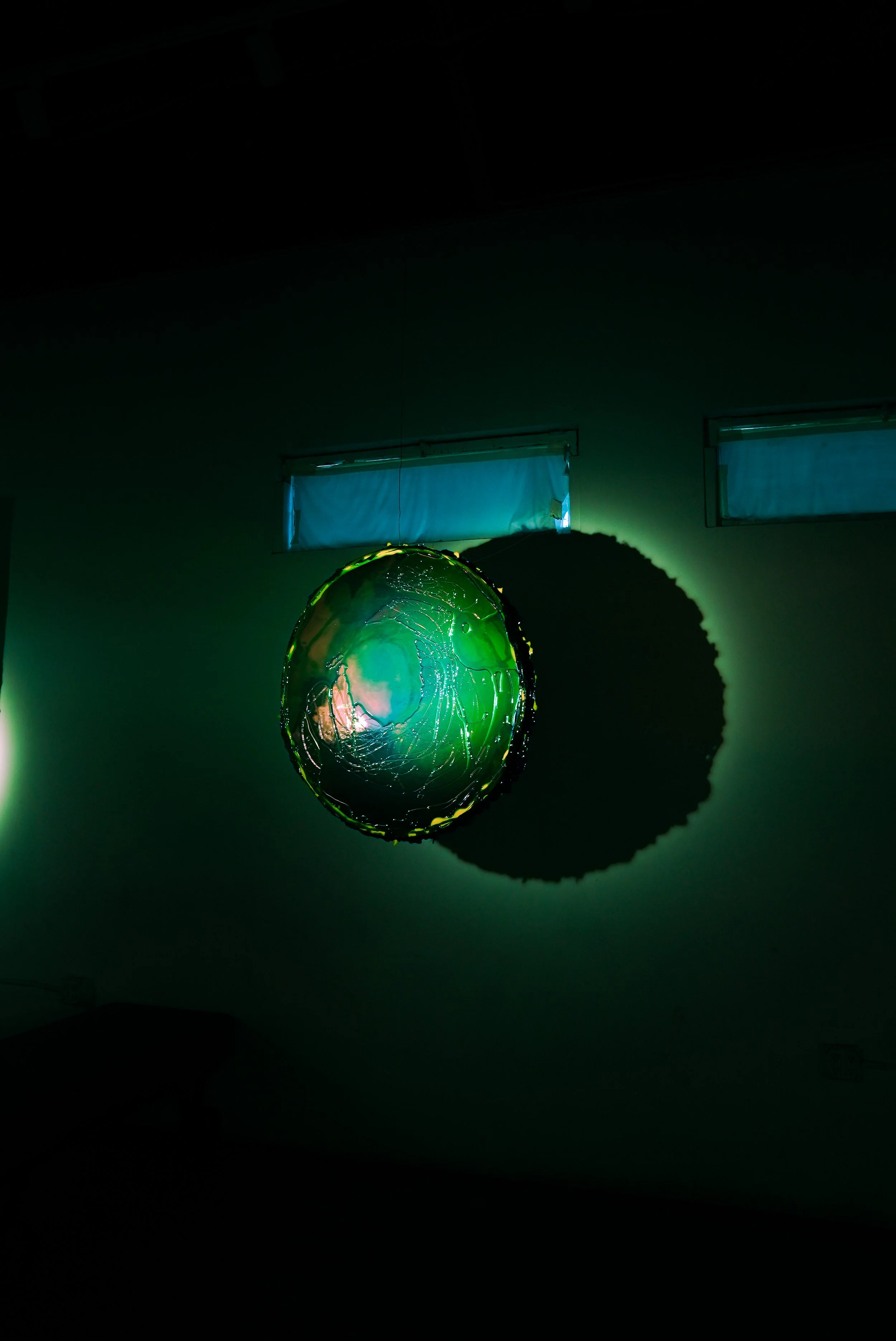 Hanging glass-like resin mirror with green and blue hues casting a shadow on the wall behind it in a dark room with small windows.