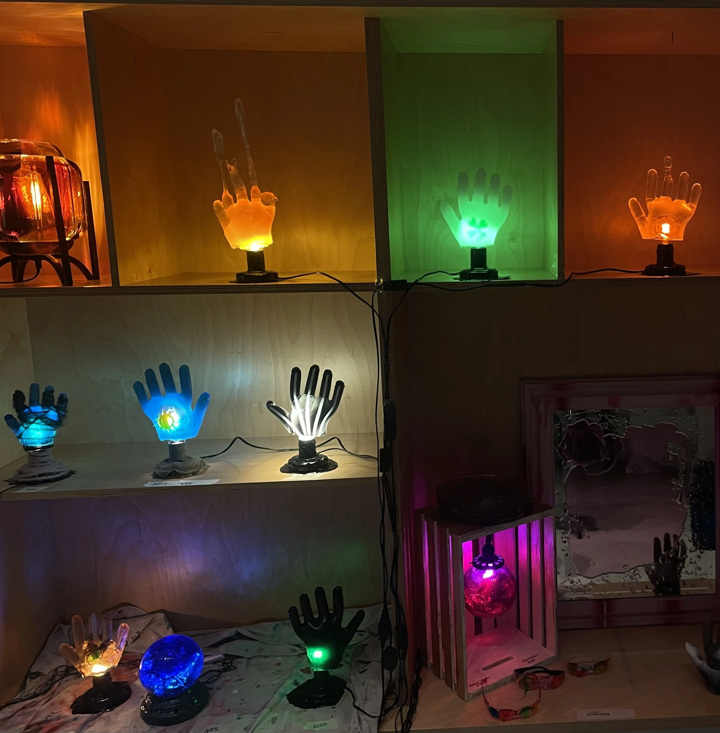Display of colorful decorative resin lamps shaped like hands and other objects, arranged on wooden shelves with some inside illuminated display cases.