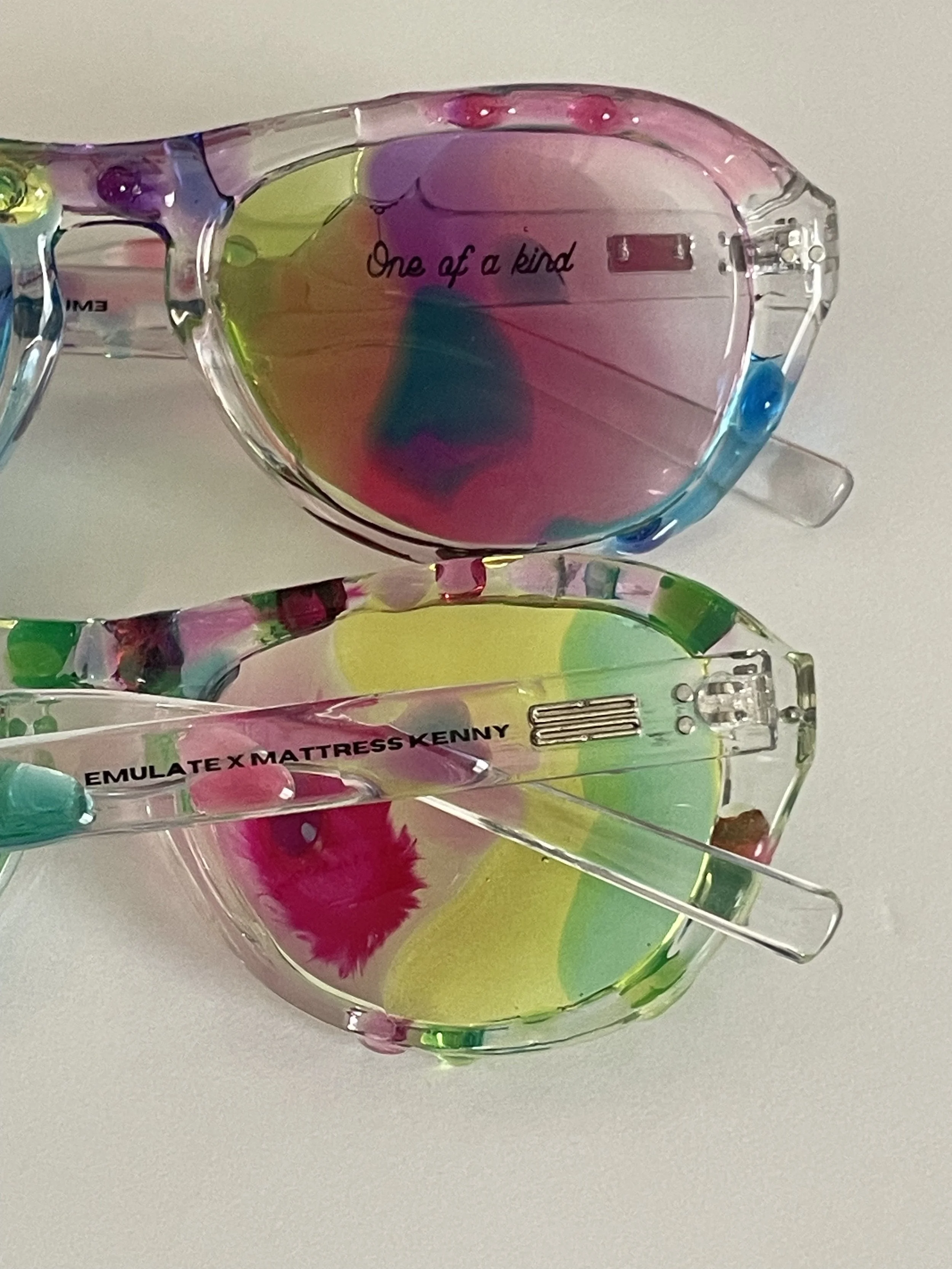 Colorful, transparent sunglasses with inscriptions that read 'One of a kind' and 'Emulate x Mattress Kenny' on the arms, resting on a white surface.