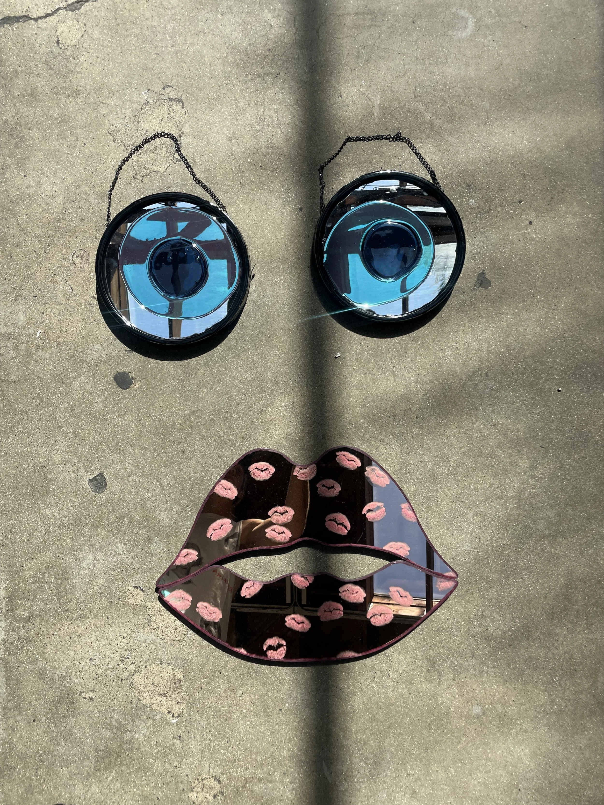 Two round decorative mirrors with chain hangers resemble eyes, and a lip-shaped mirror decorated with pink lips resembles a mouth, forming a face on the ground.