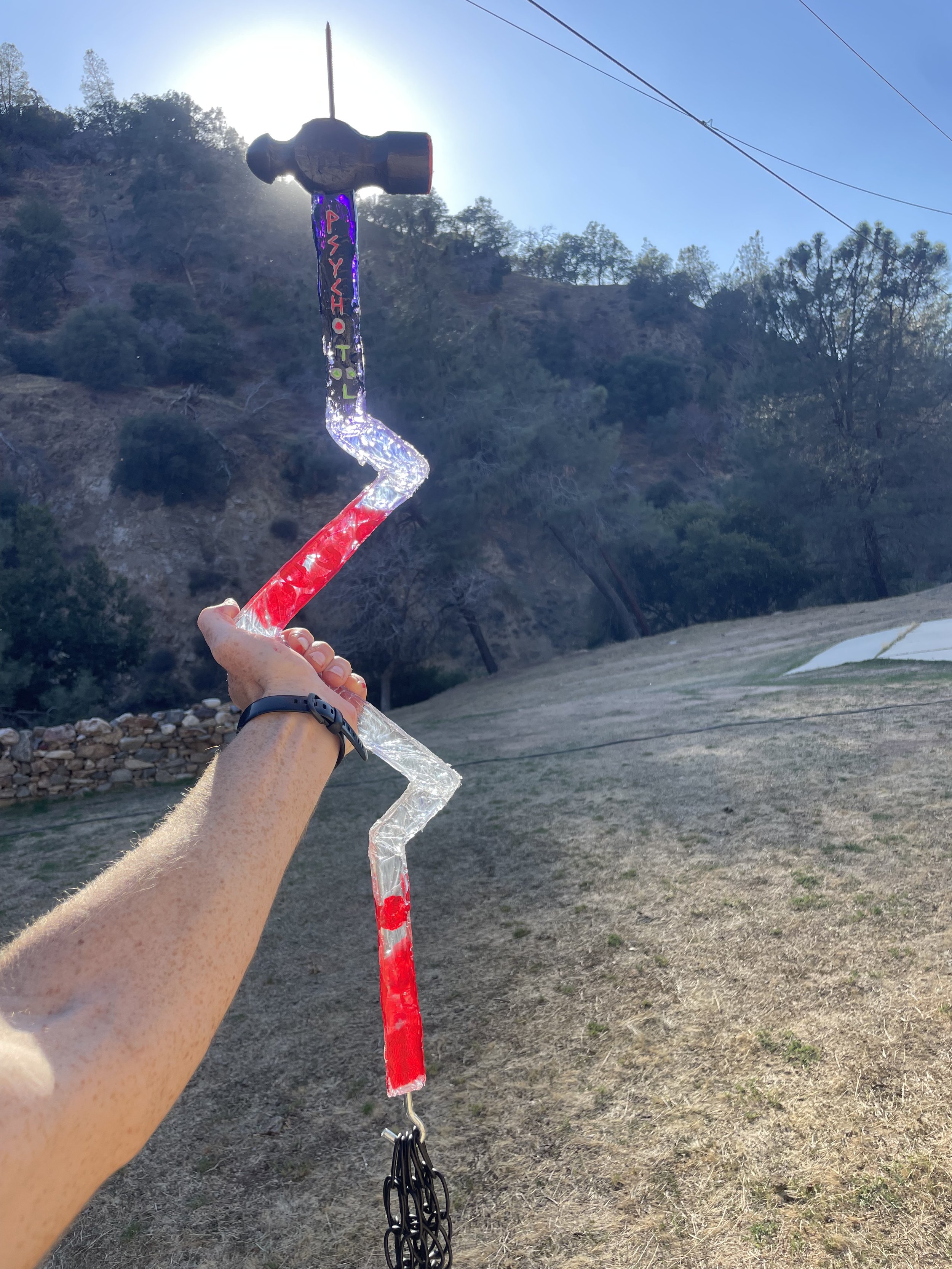 A hand holding a colorful, resin zigzag-shaped sledgehammer prop against a hillside with trees and clear blue sky.