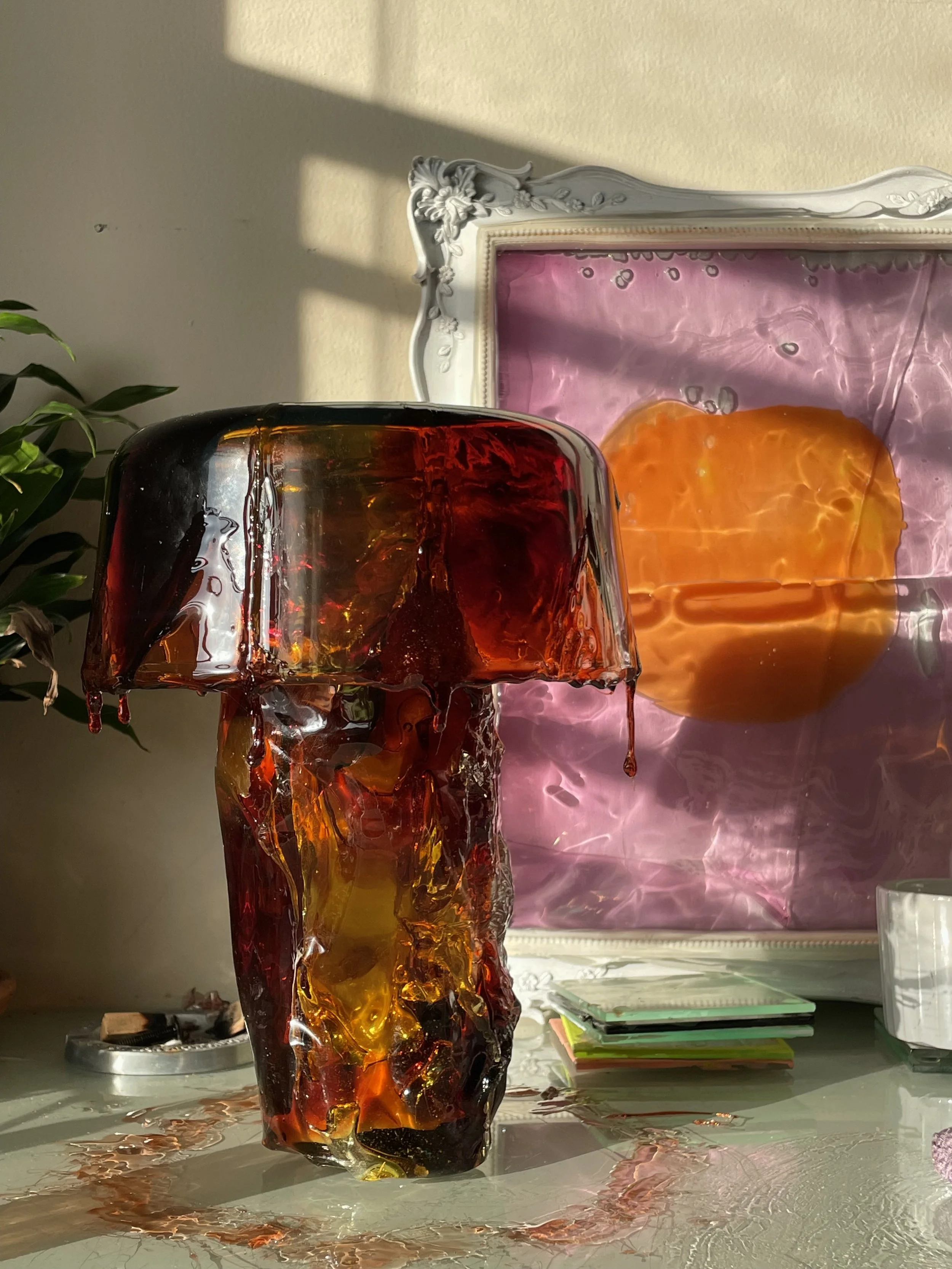 Glass and resin mushroom sculpture with a mixture of amber, red, and black colors on a reflective surface, with a pink and purple backing and a decorative white frame.