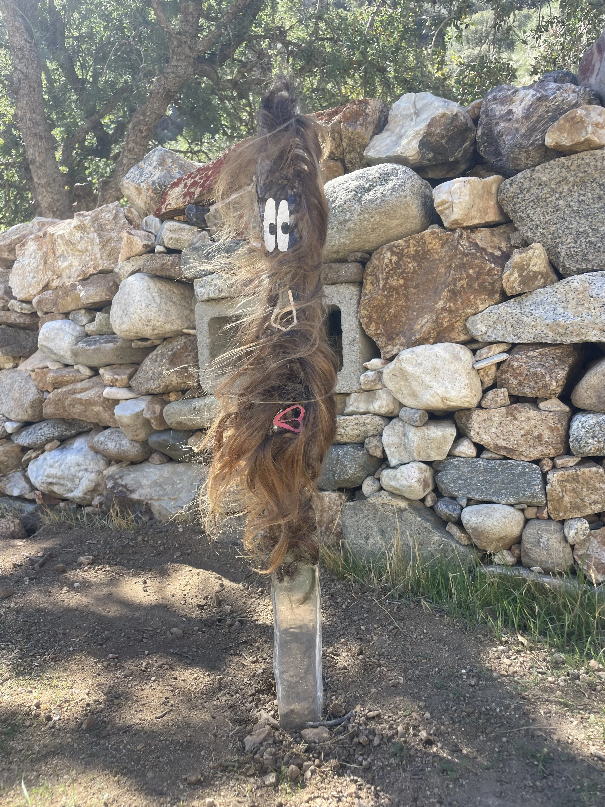 A resin plank with a face painted on it and human hair set inside, blowing in the wind. Stone wall in the background.