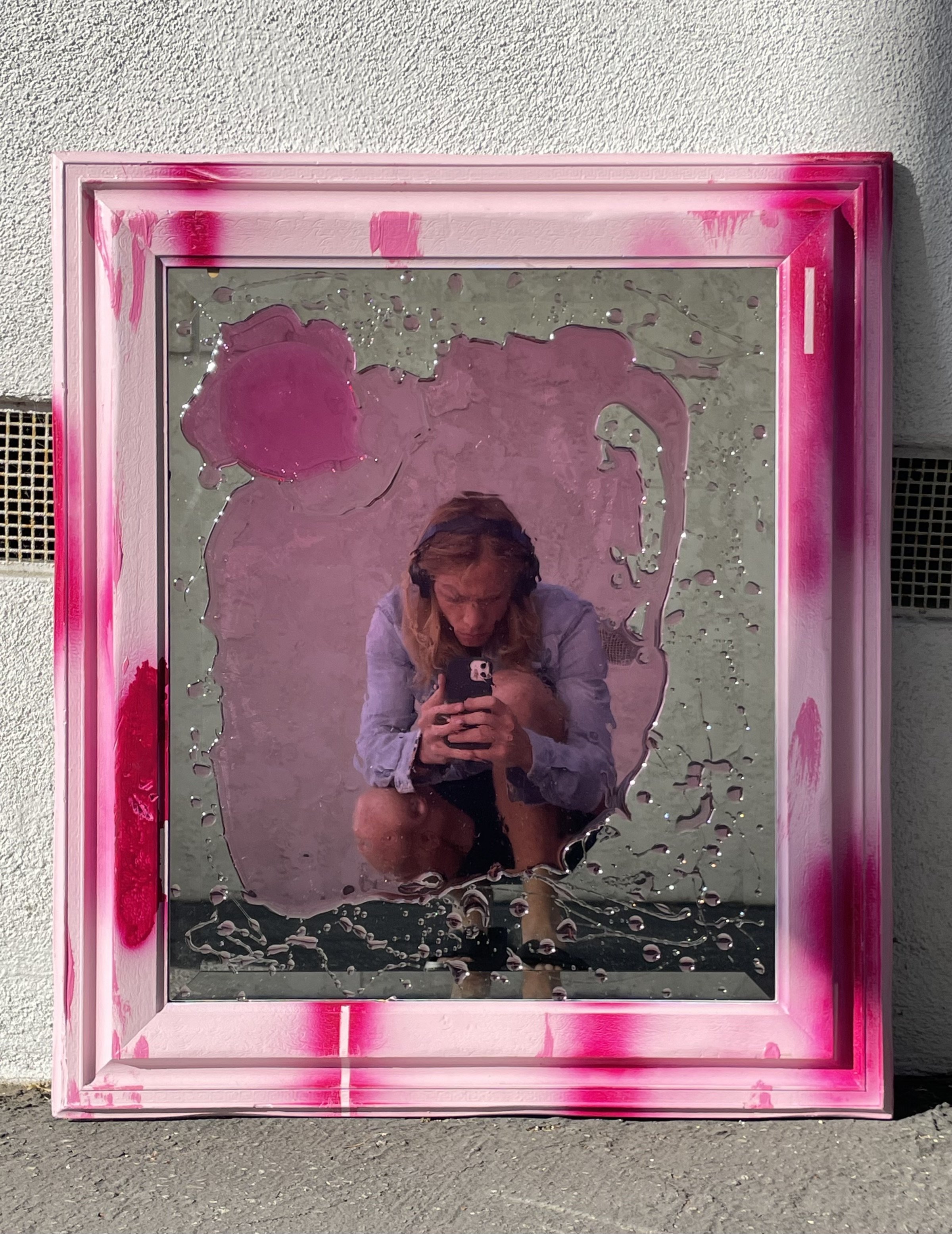 A person taking a selfie in a mirror with pink resin splattered on the edges and surface, reflecting a person with blonde hair wearing a white or light-colored top, squatting.