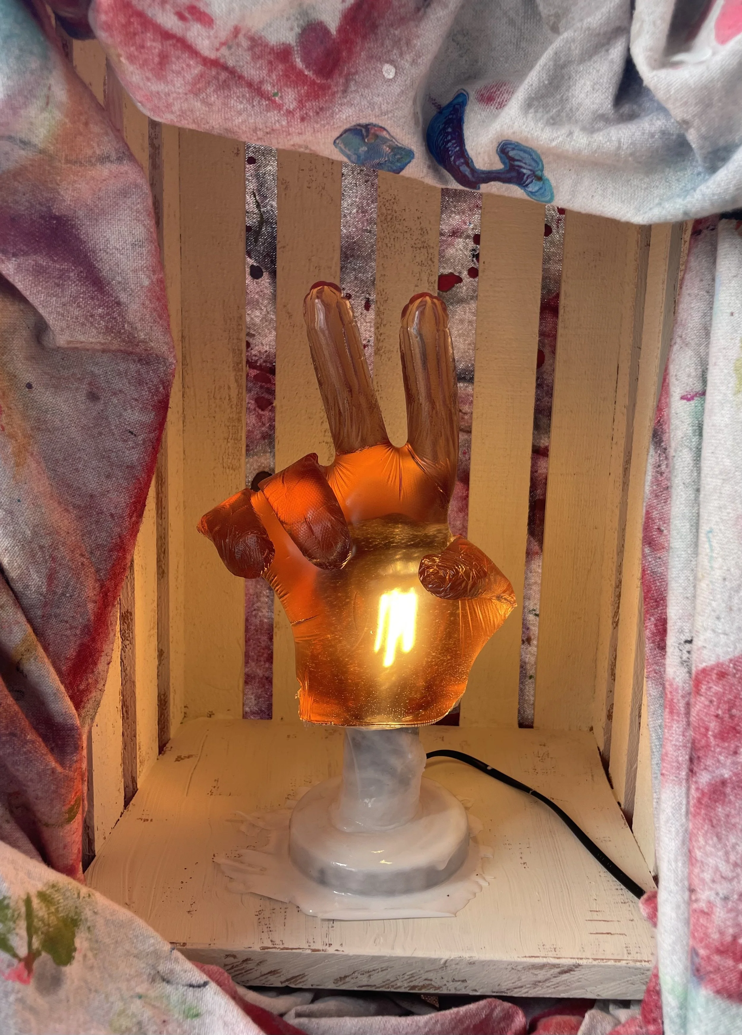 Brown glow peace sign resin hand lamp on a white base with a wooden background and colorful fabric curtains.