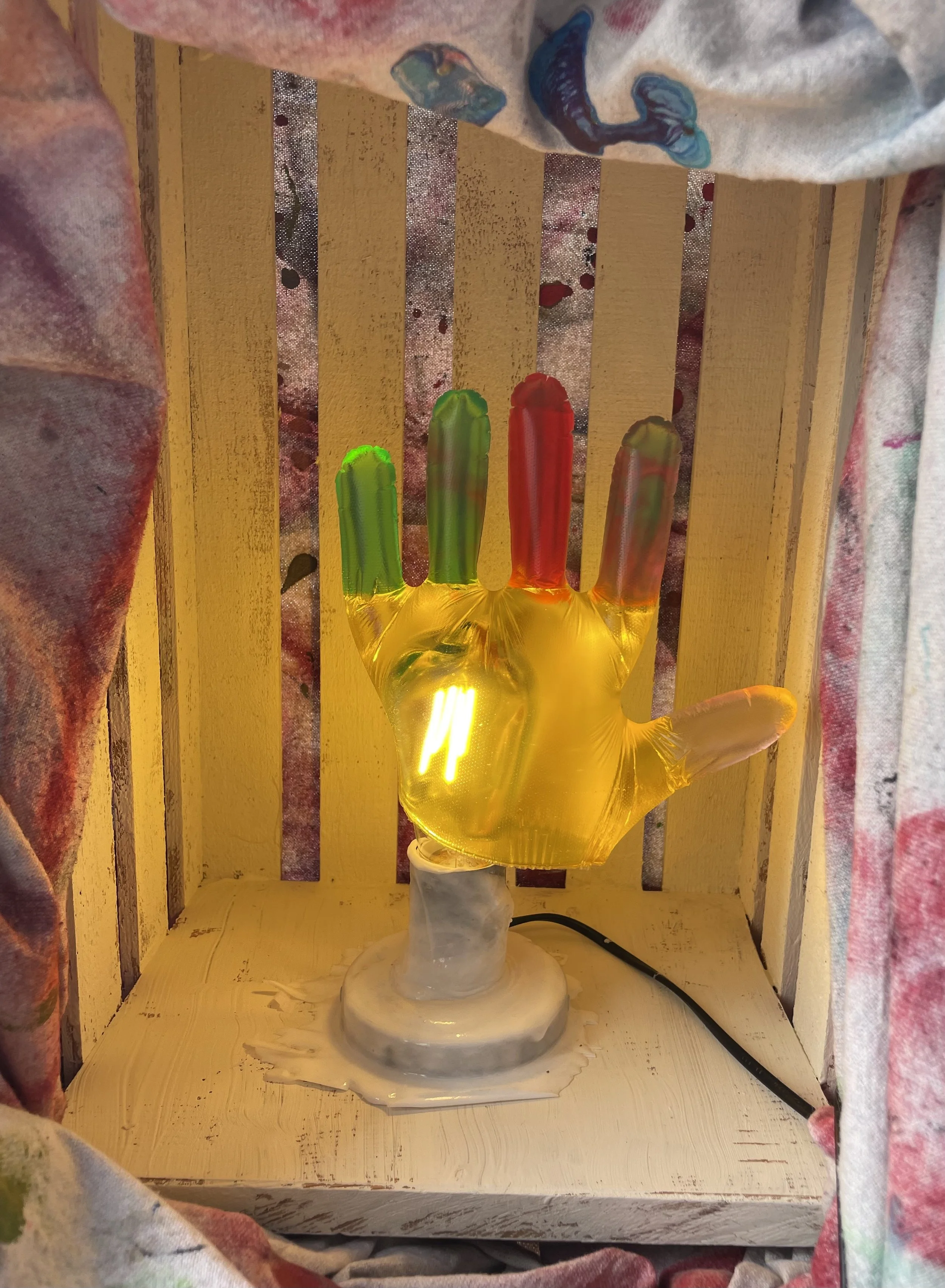 A resin sculpture of a hand with colorful fingers, illuminated from within, is displayed on a white base inside a wooden box surrounded by floral curtains.
