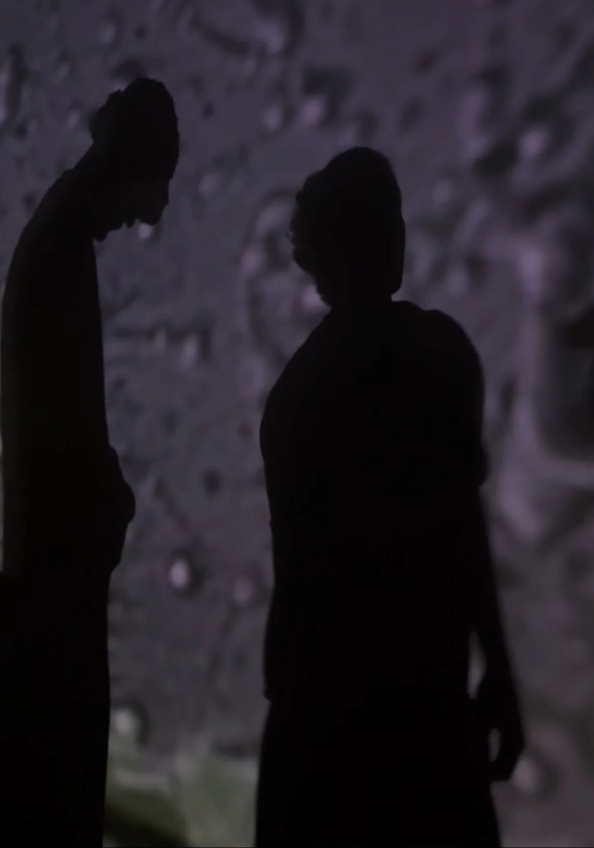 Silhouettes of a man and a woman standing close and facing each other in front of a textured, blurry background.