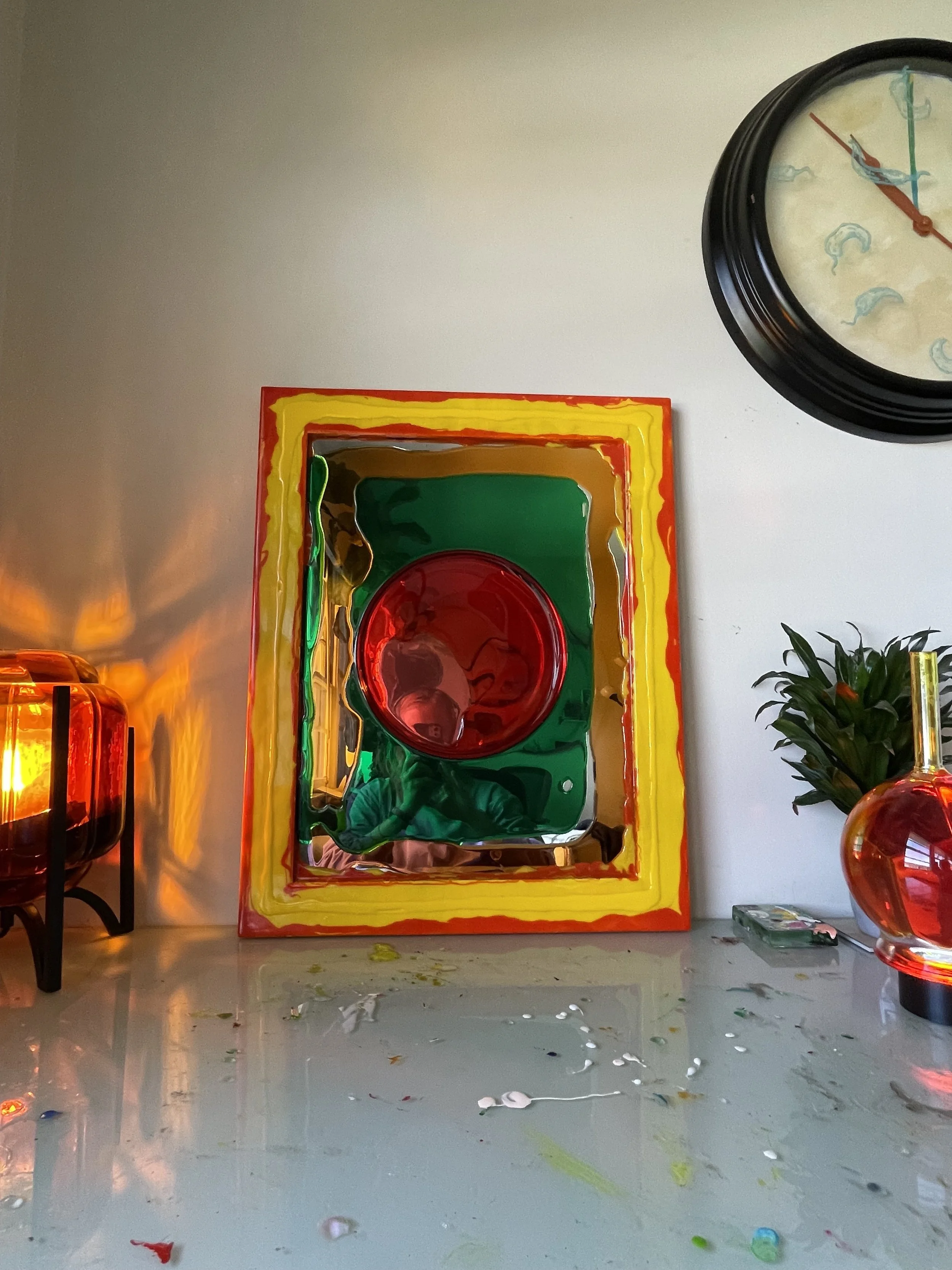 Colorful abstract resin mirror with red, green, yellow, and orange segments, placed on a reflective surface, with a decorative red resin and glass object and a plant to the side.