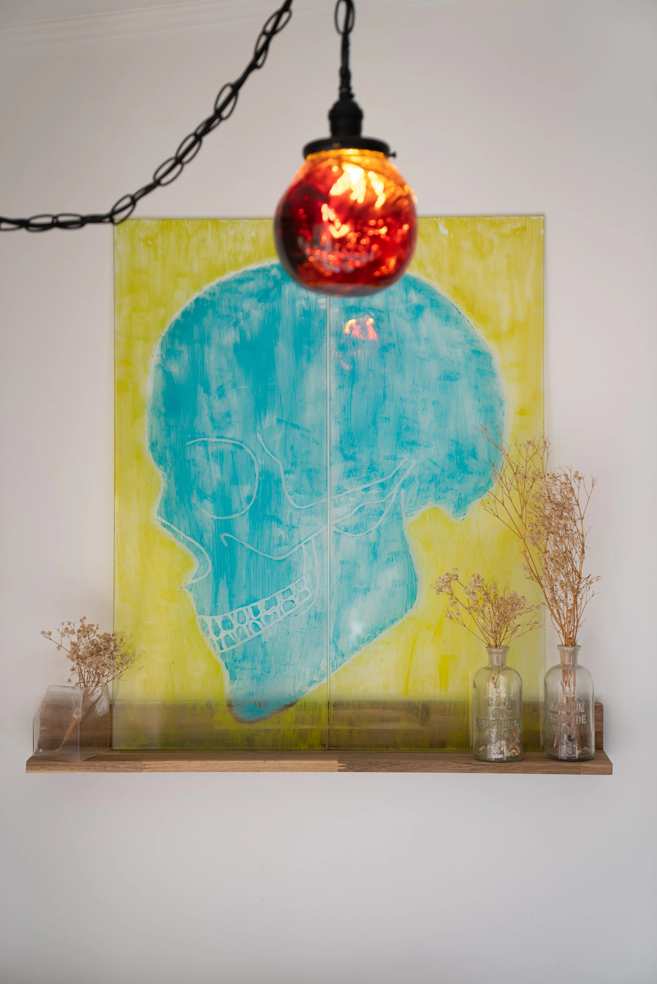 A wall-mounted wooden shelf with dried flowers in glass bottles, a colorful abstract painting of a blue skull on a yellow background, and a red pendant resin light hanging above.