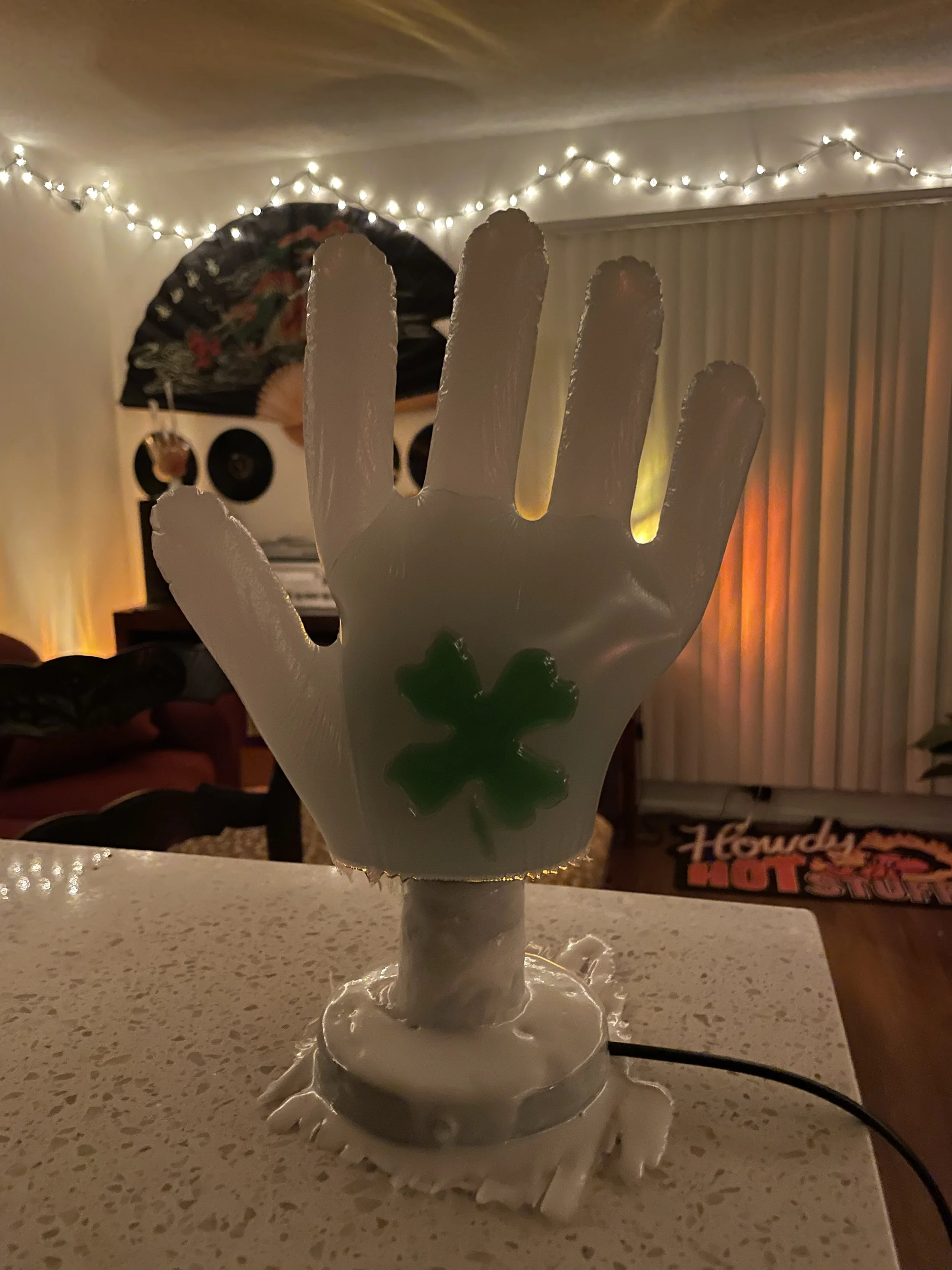 A white resin lamp shaped like a human hand with a green shamrock on the palm, placed on a table in a room decorated with string lights and wall art.