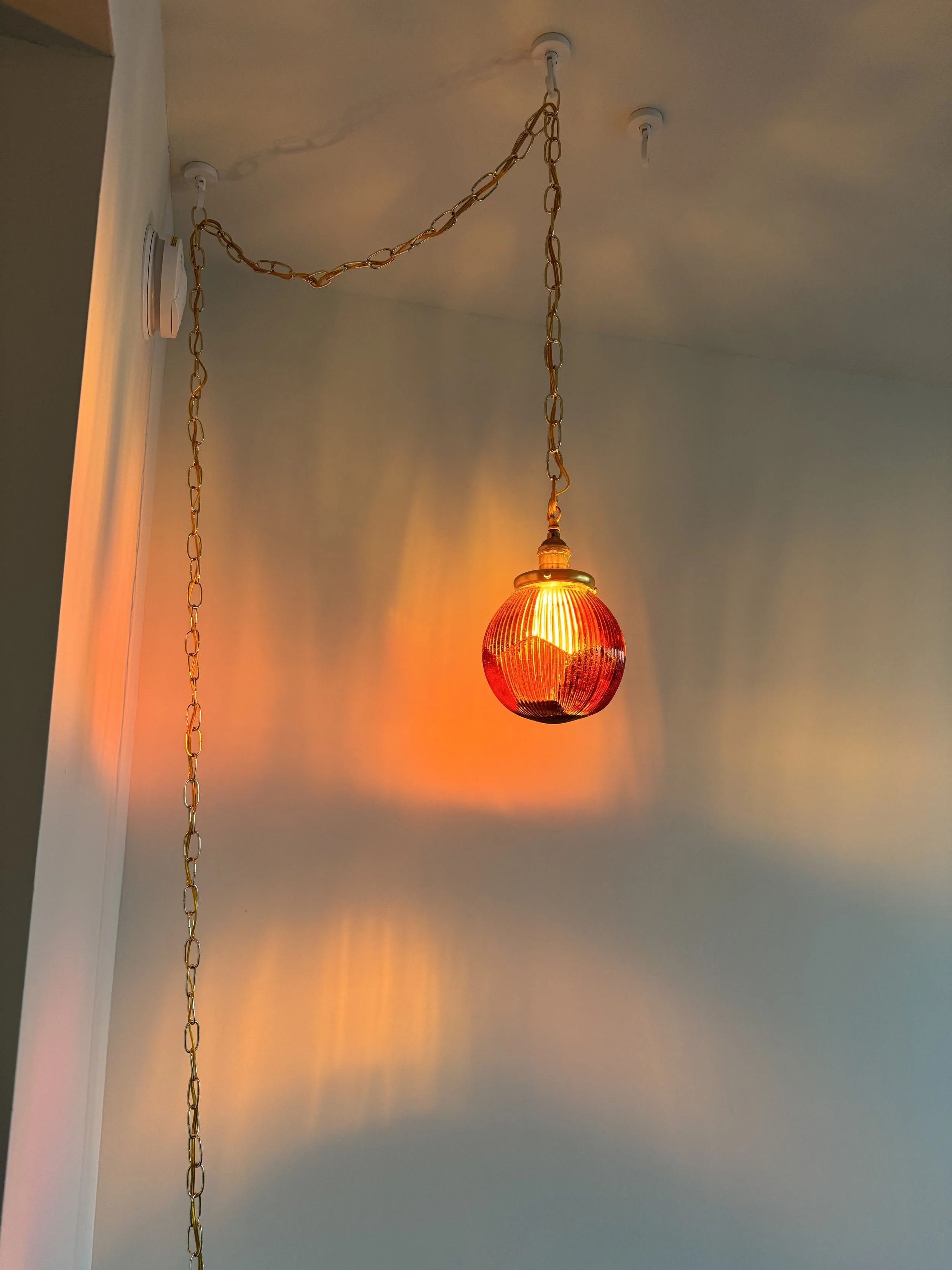 An orange-red pendant resin light fixture hanging from the ceiling with a gold chain. The light casts colorful reflections on the wall.