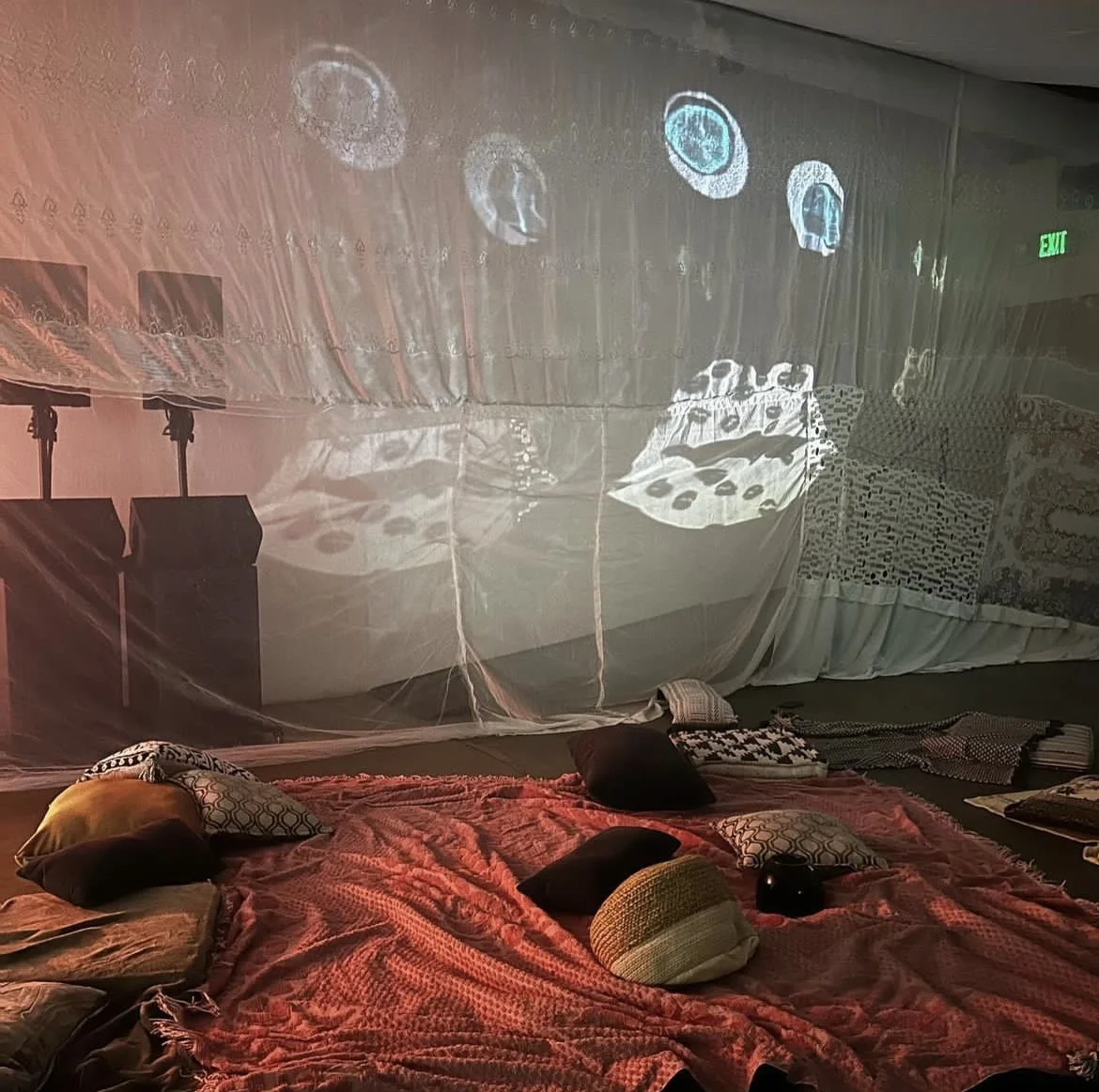 A cozy scene with pillows and a blanket on the floor, with a sheer curtain decorated with the reflection of resin mirrors to resemble a face