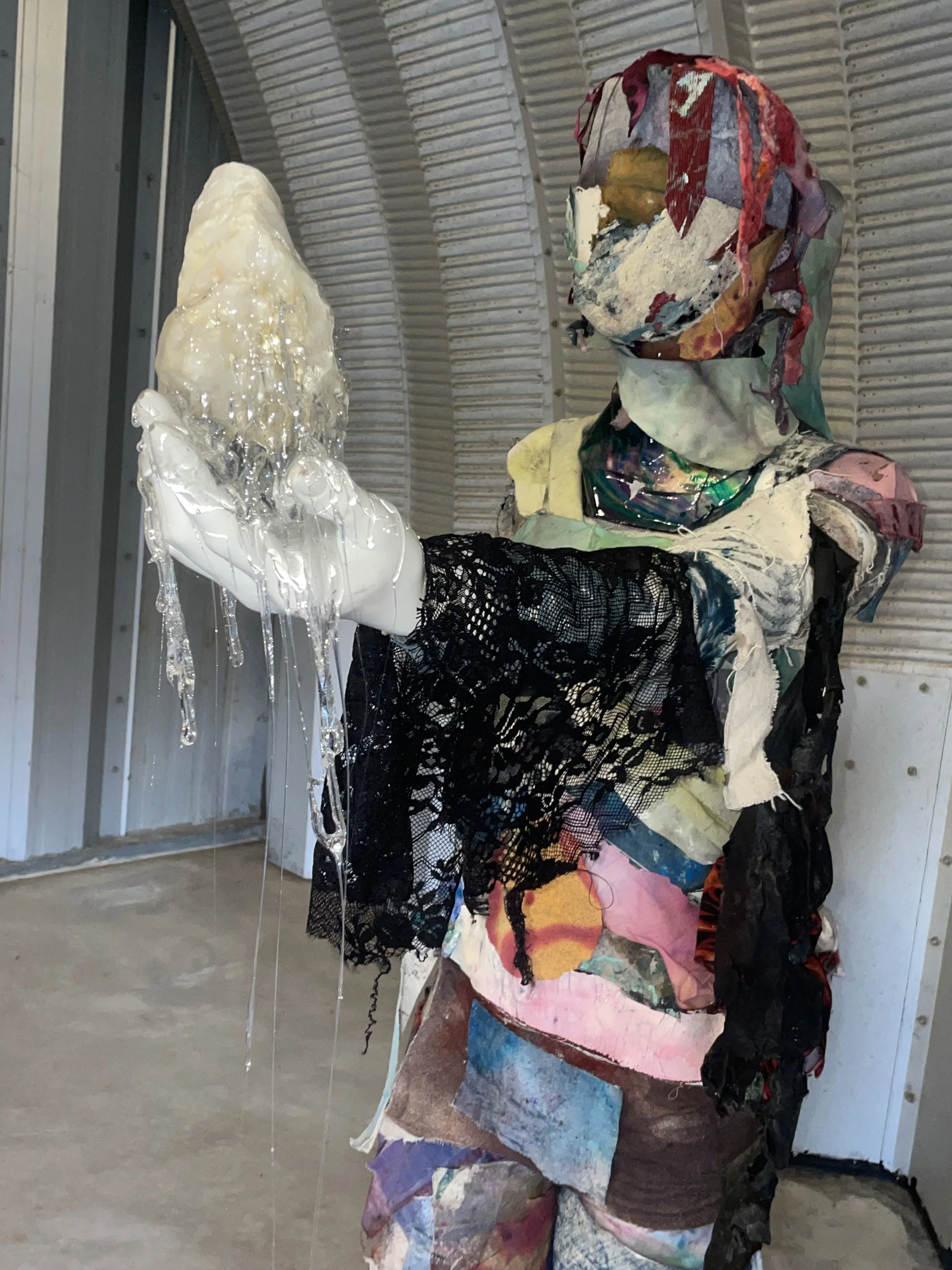 An abstract sculpture resembling a human figure, made from colorful fabric and mixed materials, holding a large quartz crystal being suspended in clear resin.