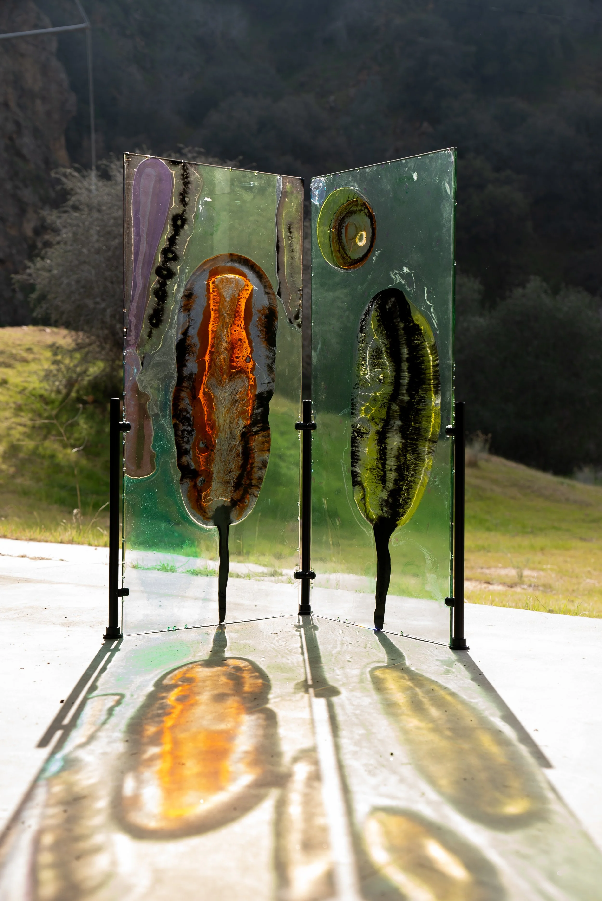 Resin art installation with two panels featuring abstract shapes and colors, casting reflections on the ground outdoors with a mountainous background.