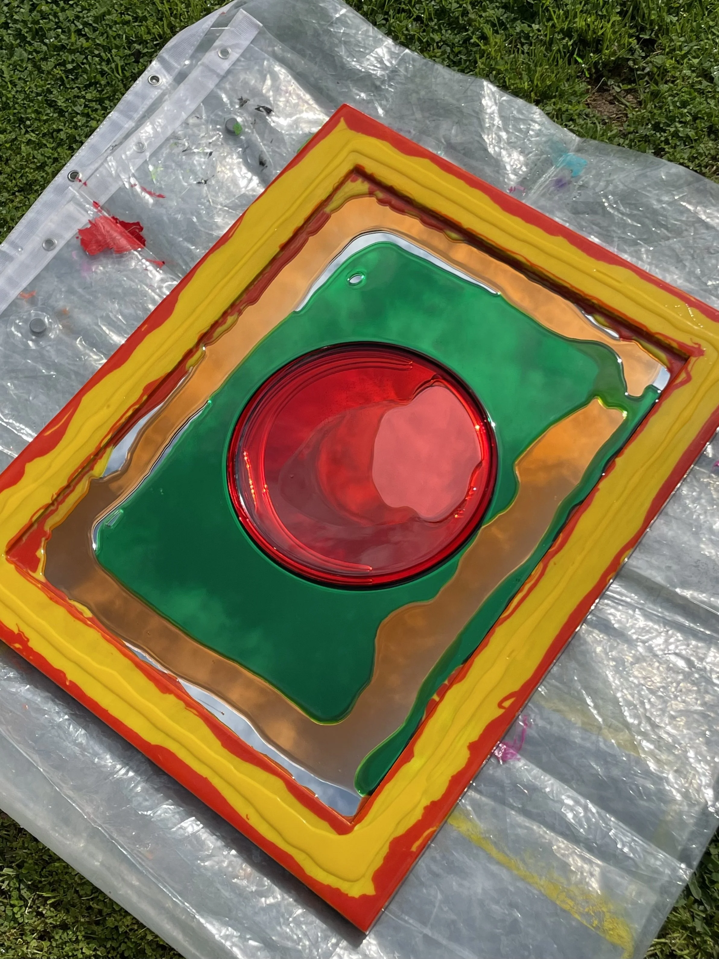 Colorful resin artwork with a red circular center, surrounded by green, yellow, orange, and red borders, placed on a reflective surface outdoors.