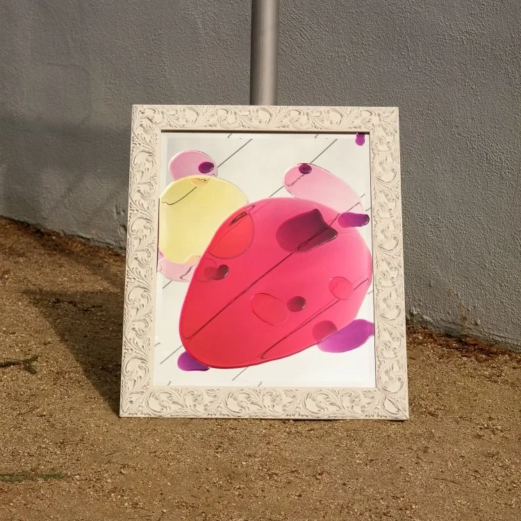 Colorful resin mirror displayed in a decorative white frame, leaning against a gray wall on a dirt ground.