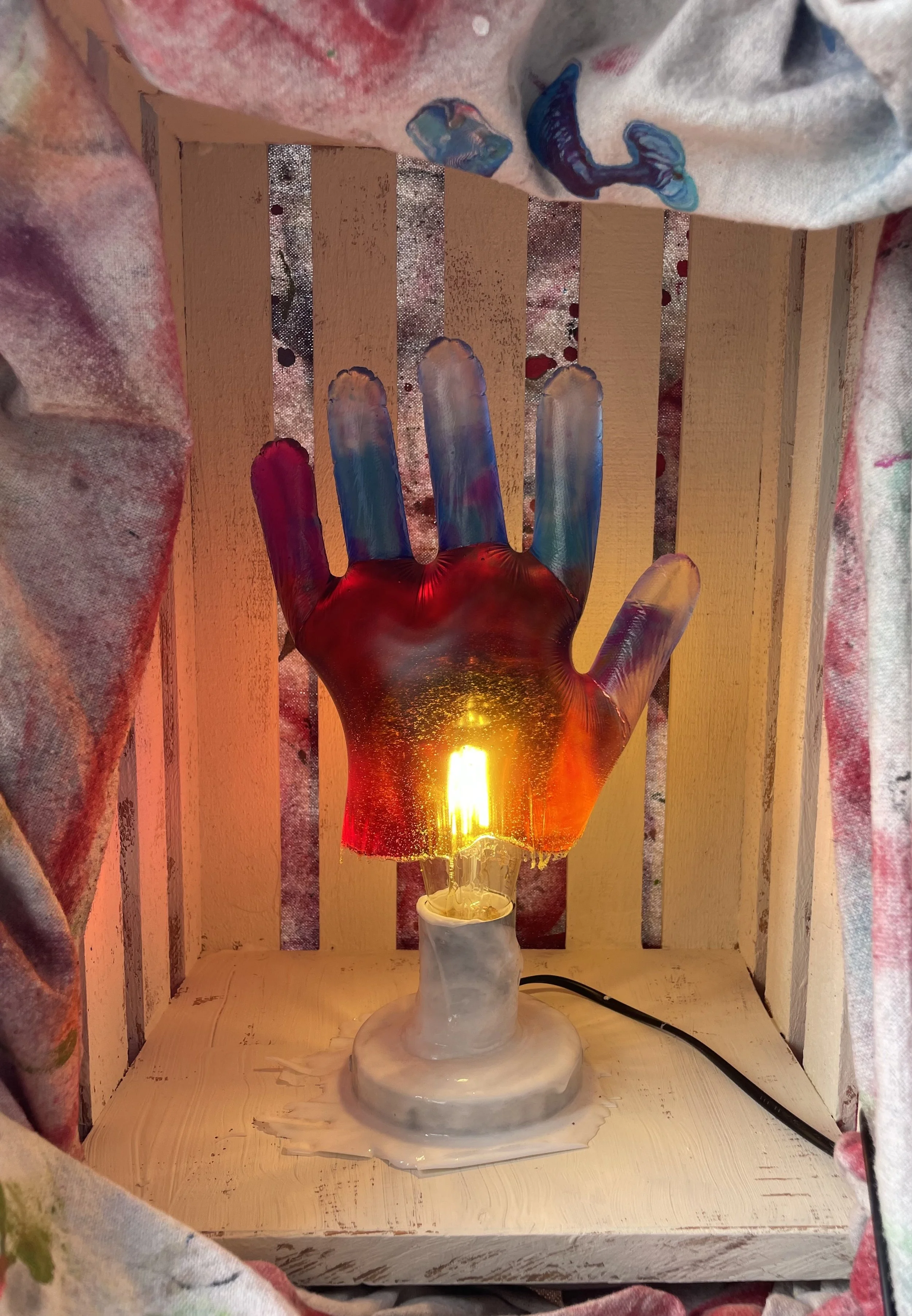 A decorative hand-shaped resin lamp with colored fingers, placed on a wooden surface within a small wooden frame, glowing warmly with a yellow-orange light.