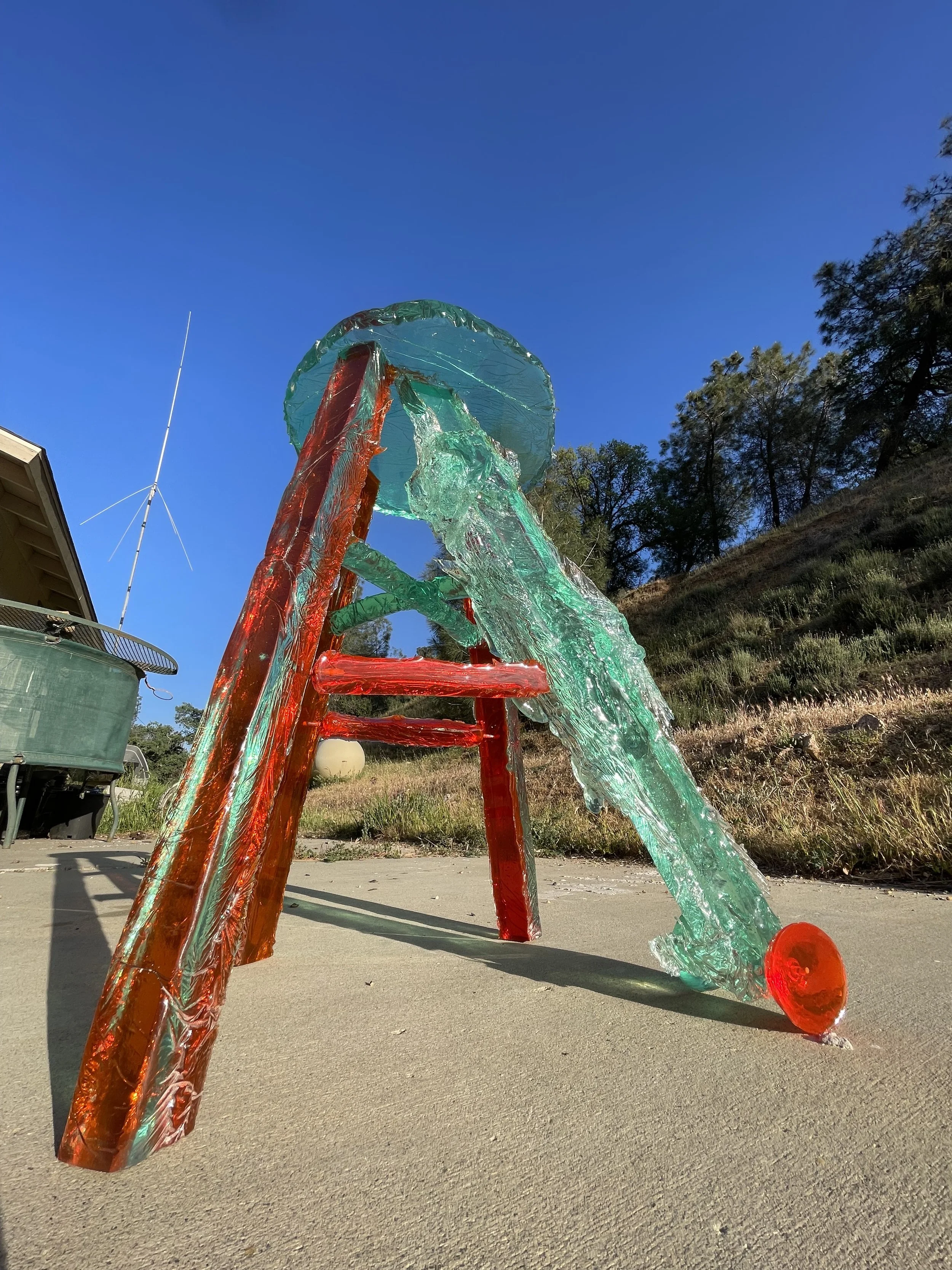 Glass sculpture of a slide, made of colorful glass with red and turquoise hues, outdoors under a clear blue sky.