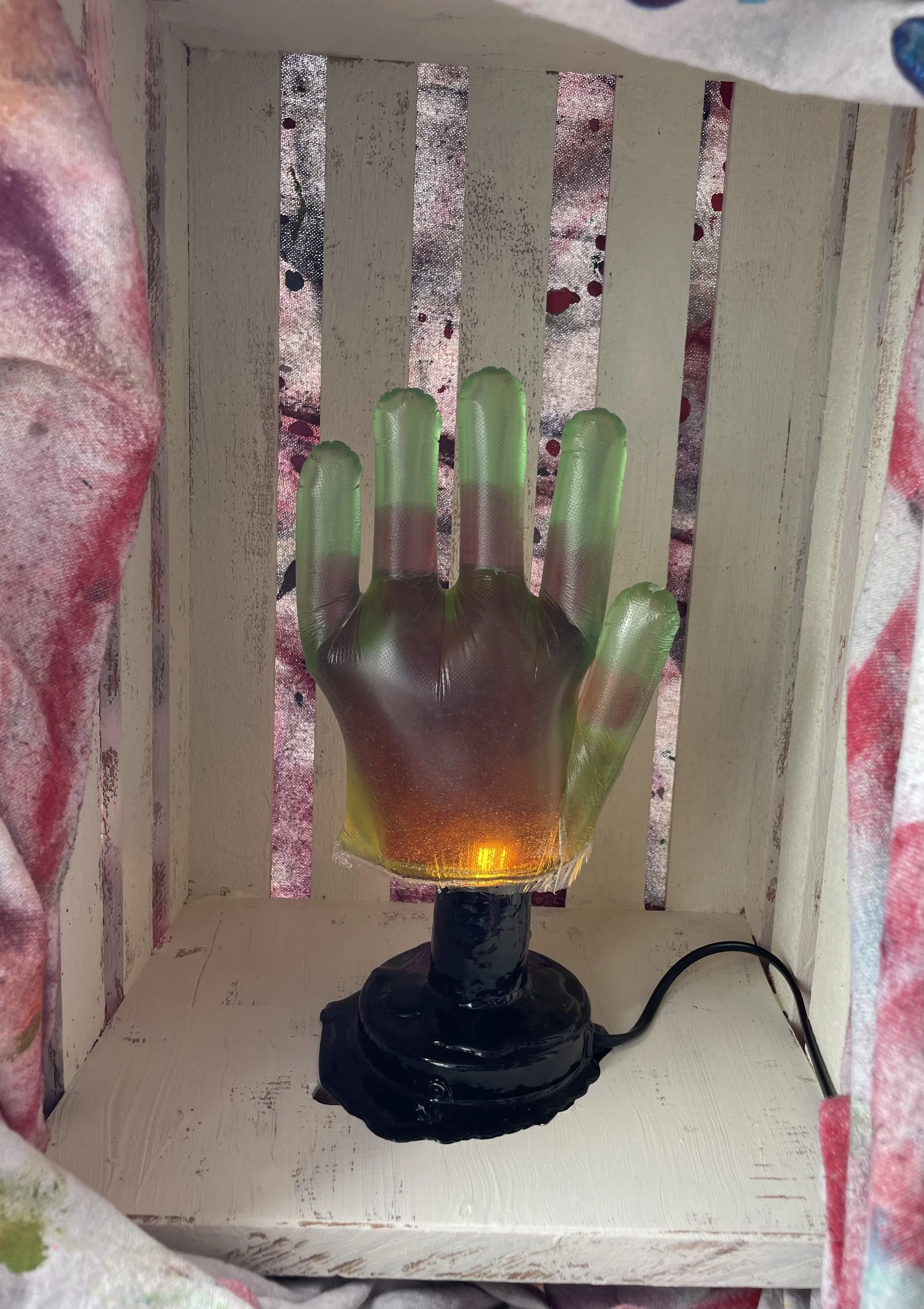 A decorative lamp shaped like a hand with fingers pointing upward, made of colorful resin with shades of green, pink, and amber, on a black base, placed inside a wooden box with vertical slats.