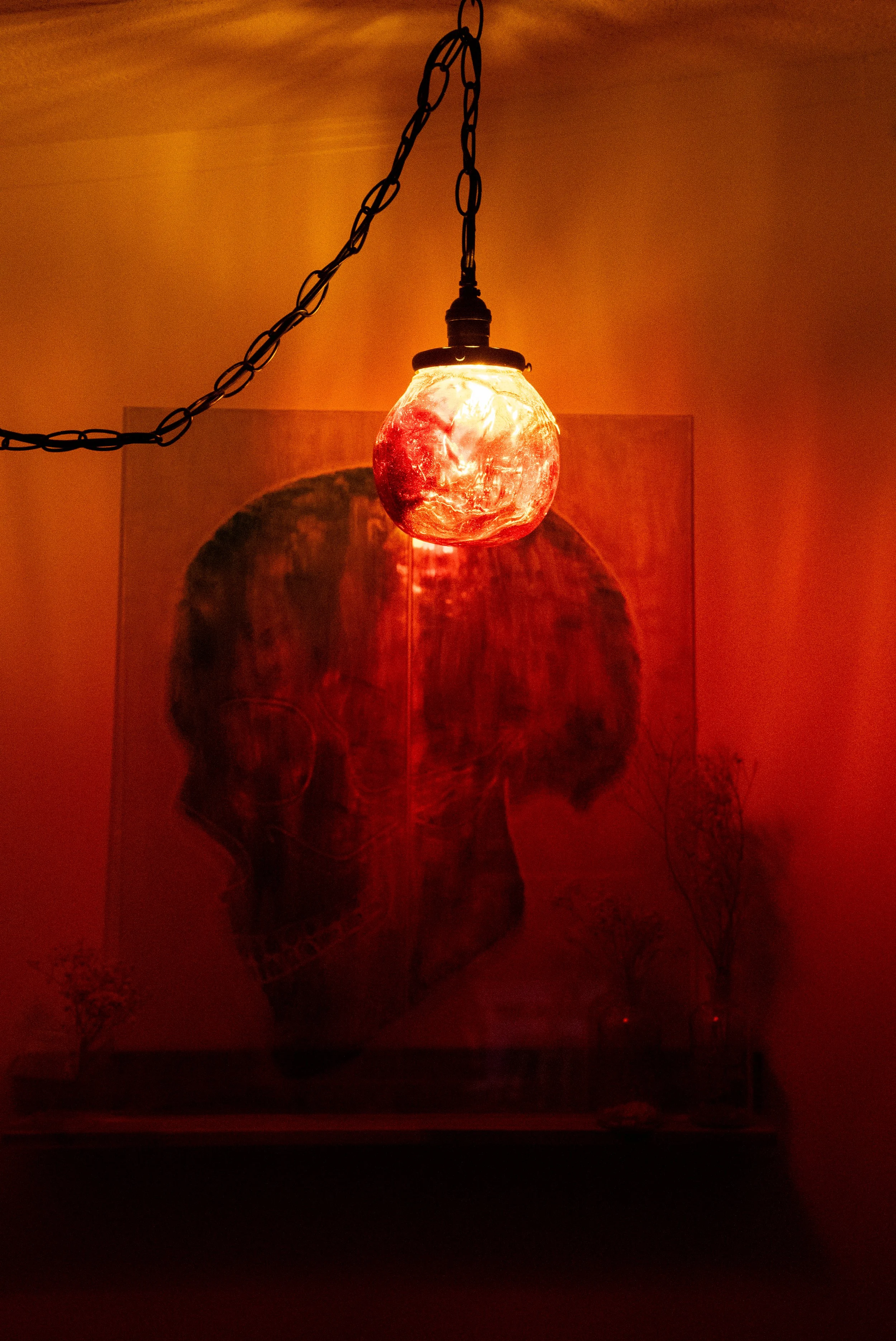A ceiling lamp made of a red, textured resin orb, casting a red glow and shadow on the wall behind, with a painting of a skull and minimal decor below.