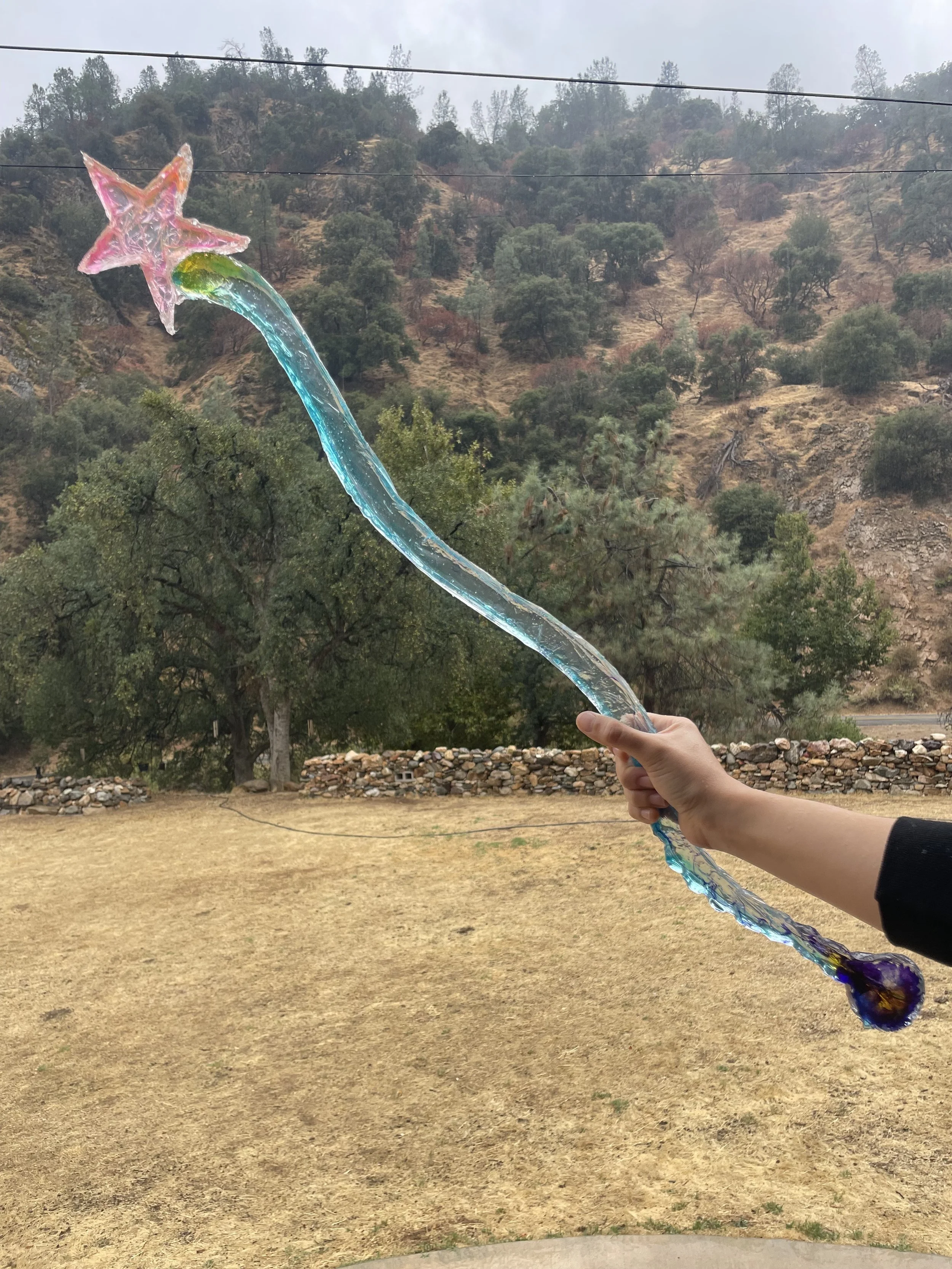 A person holding a colorful star-shaped magic resin wand with a trail, outdoors in front of a hilly landscape with trees and rocks.