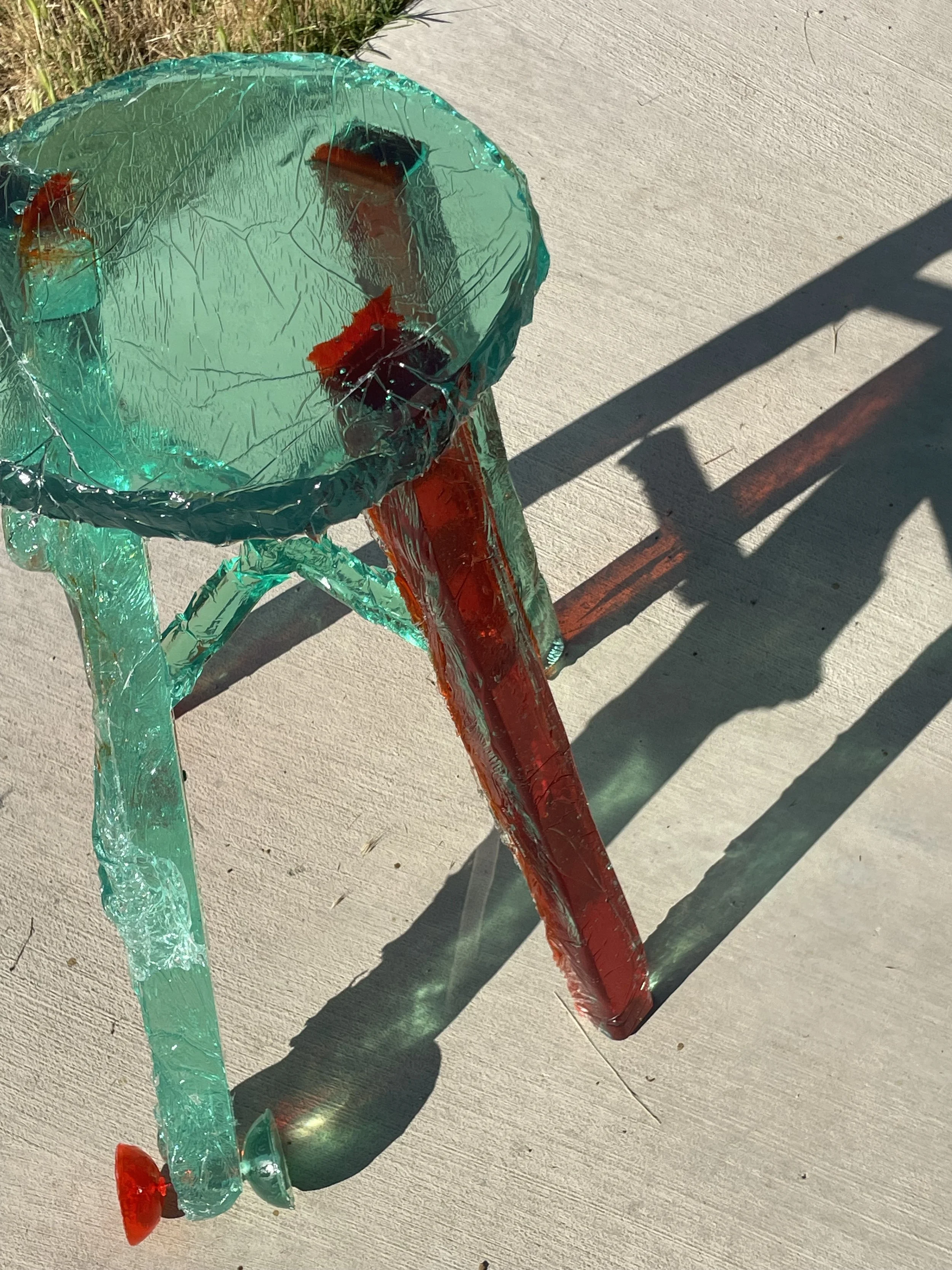 A small chair made of transparent plastic with red accents, casting colorful shadows on the concrete ground outside in sunlight.