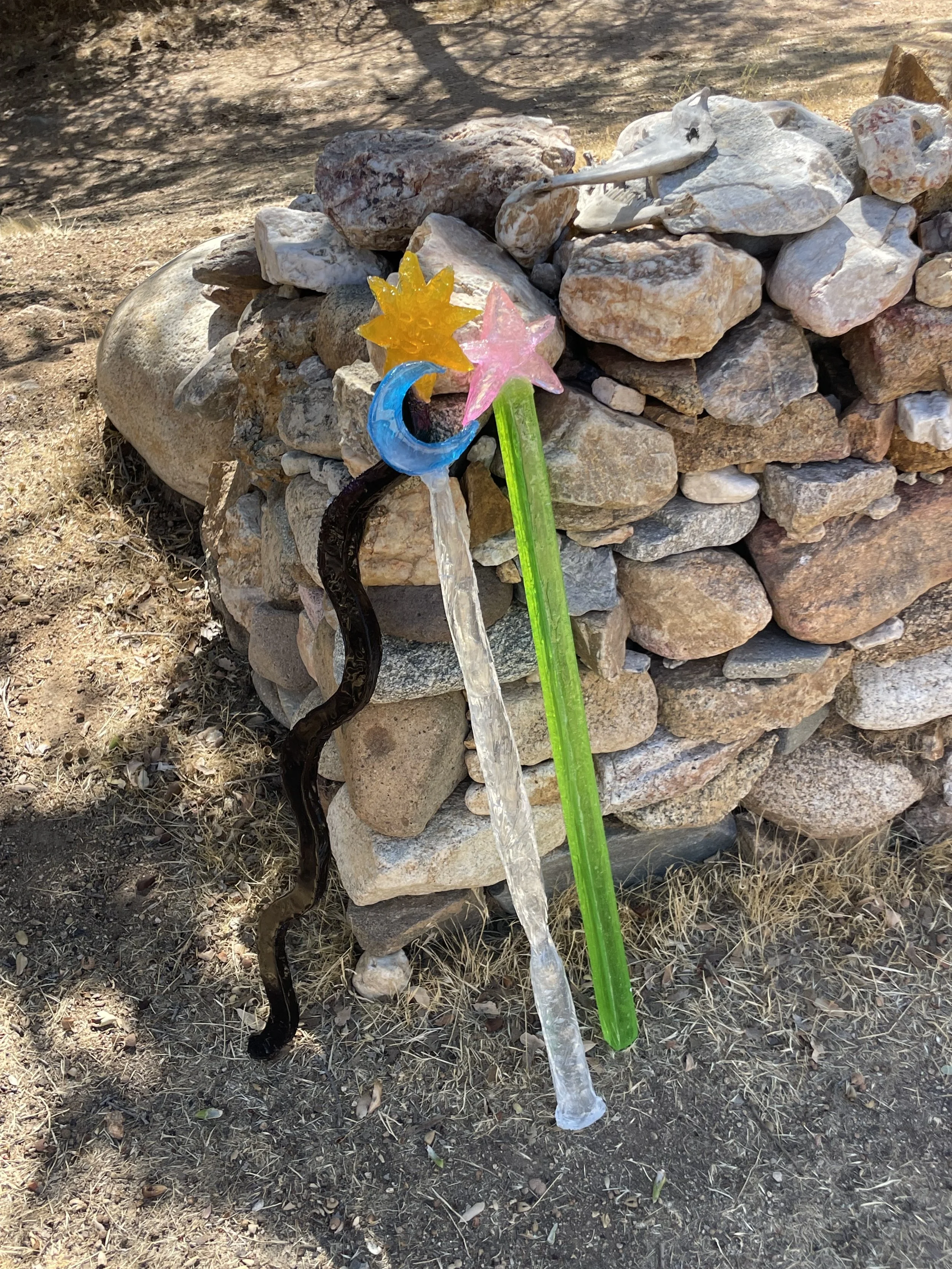 Three colorful resin fairy wands leaning against a small stone wall outdoors with dry grass and dirt ground.