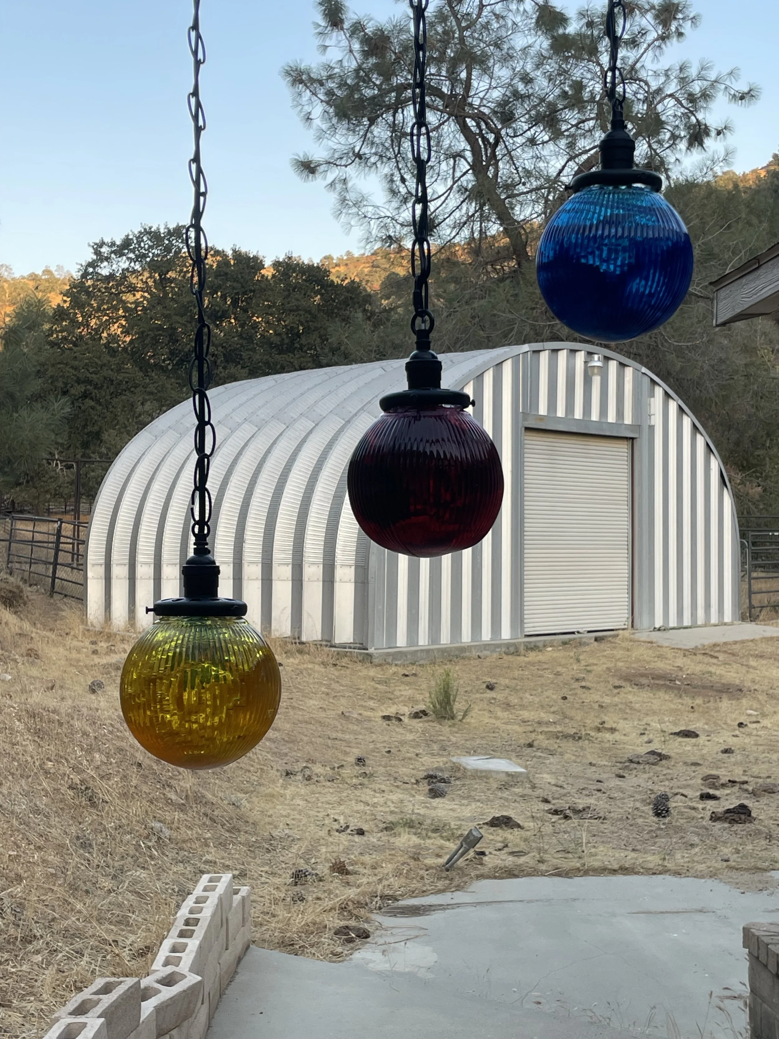 Three colored resin pendants hanging outdoors in front of a metal shed with a roll-up door, with trees and hills in the background.