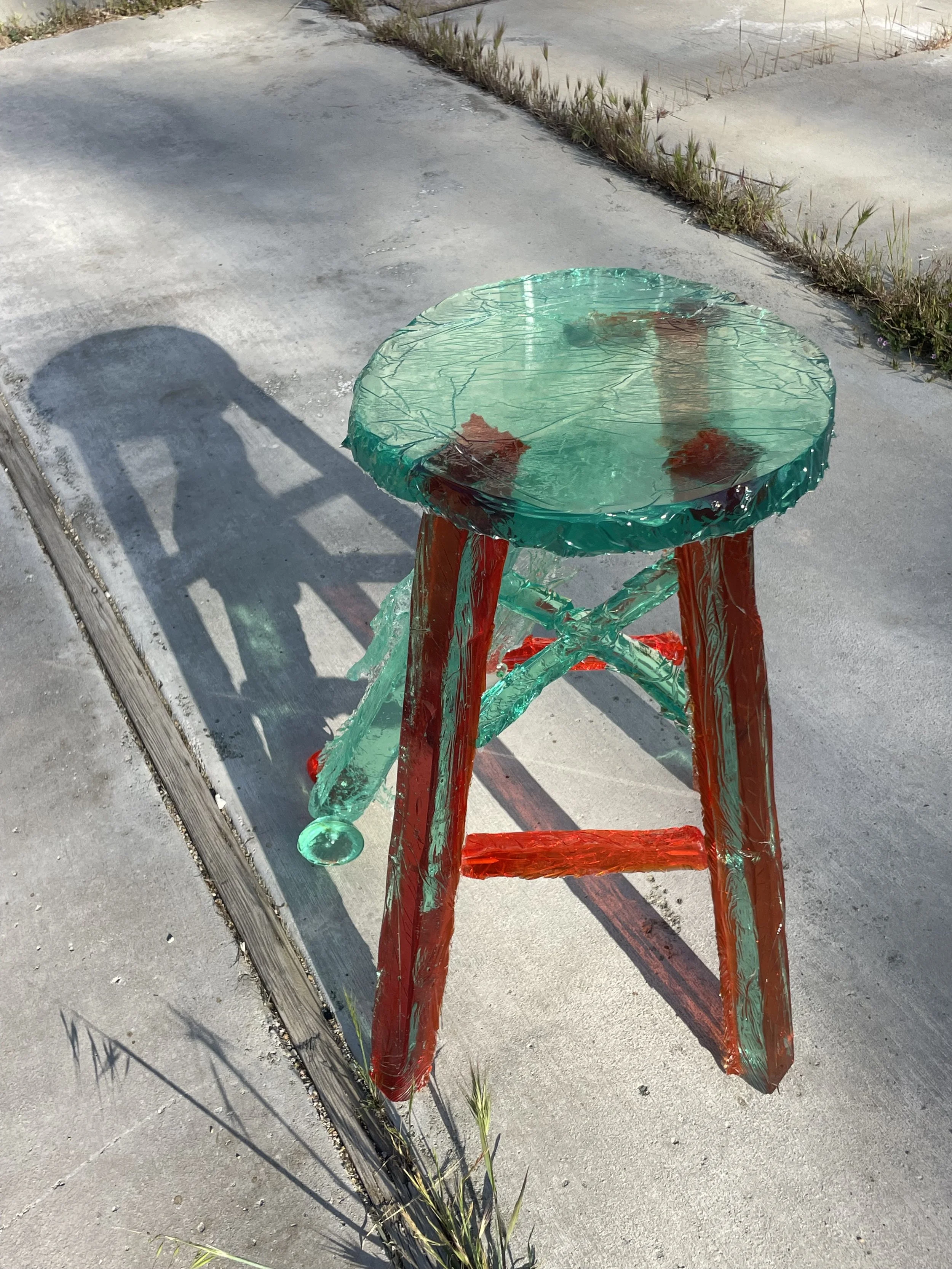 A glass-like resin stool with four legs, made of translucent green and red glass, seated on a concrete sidewalk with grass and weeds nearby.