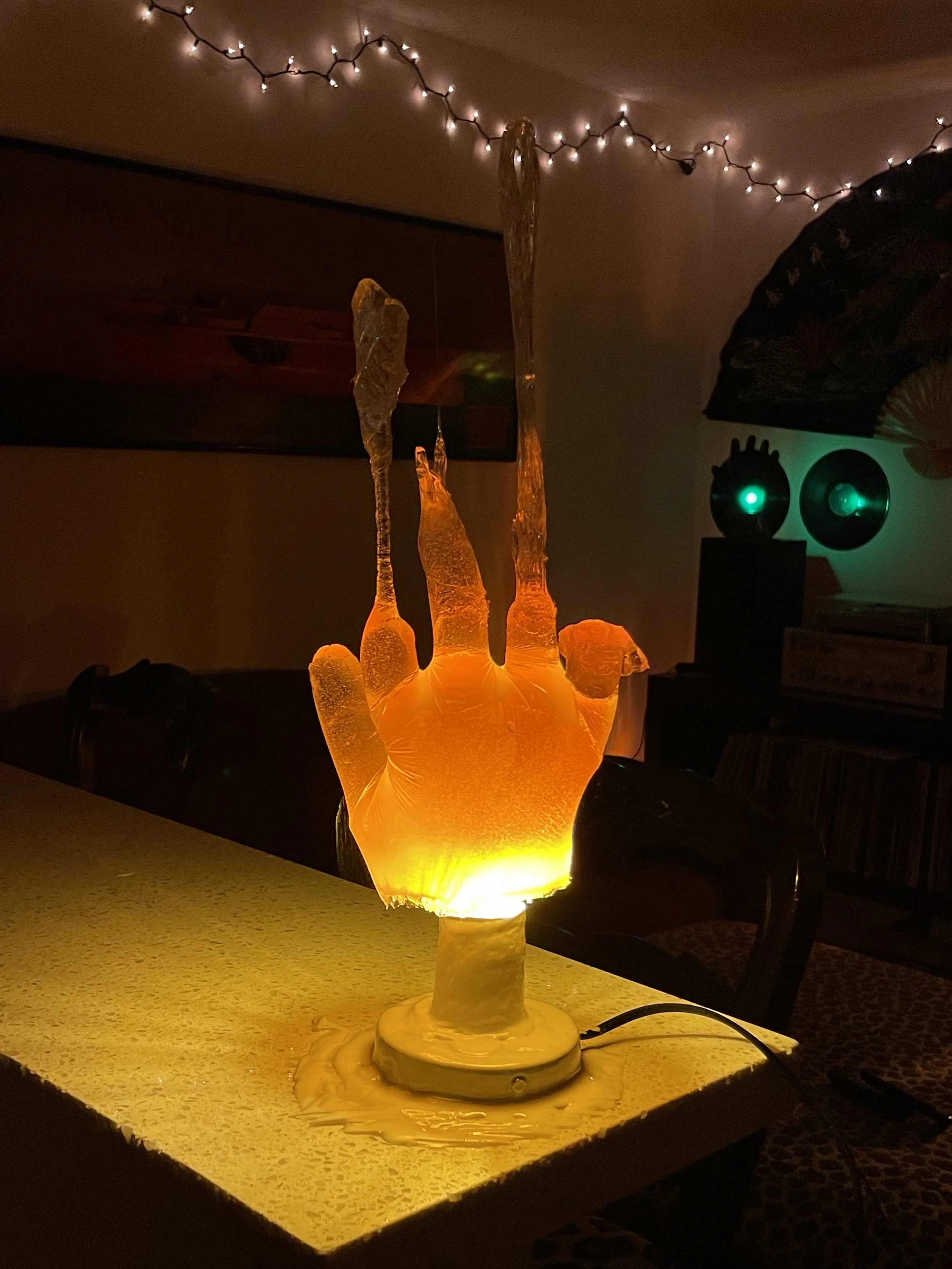 A glowing resin lamp in the shape of a hand with fingers raised, emitting an orange light, placed on a table in a dimly lit room decorated with string lights and wall art.
