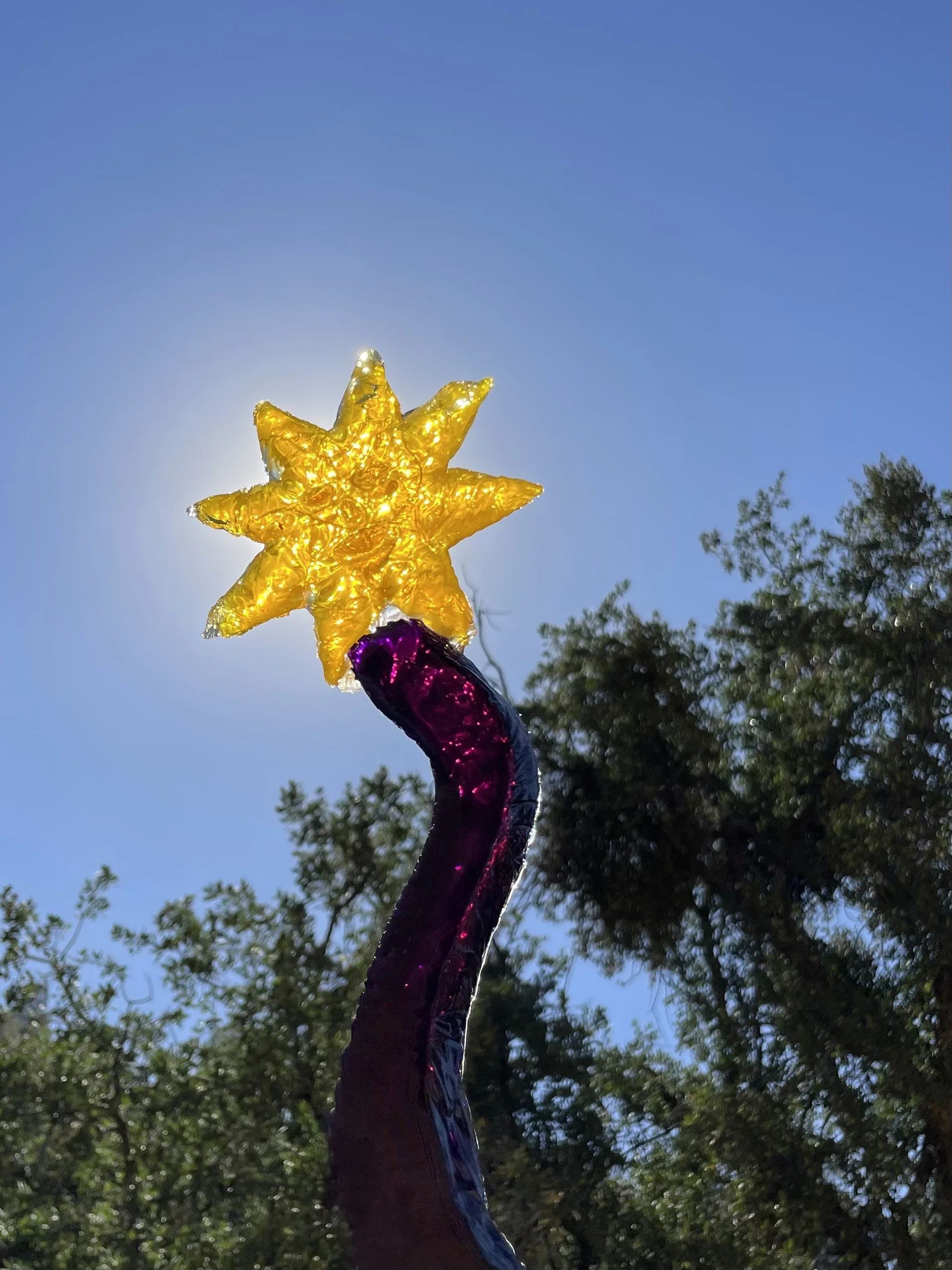 A yellow resin sun at the top of a purple resin magic wand silhouetted against a blue sky with trees in the background.