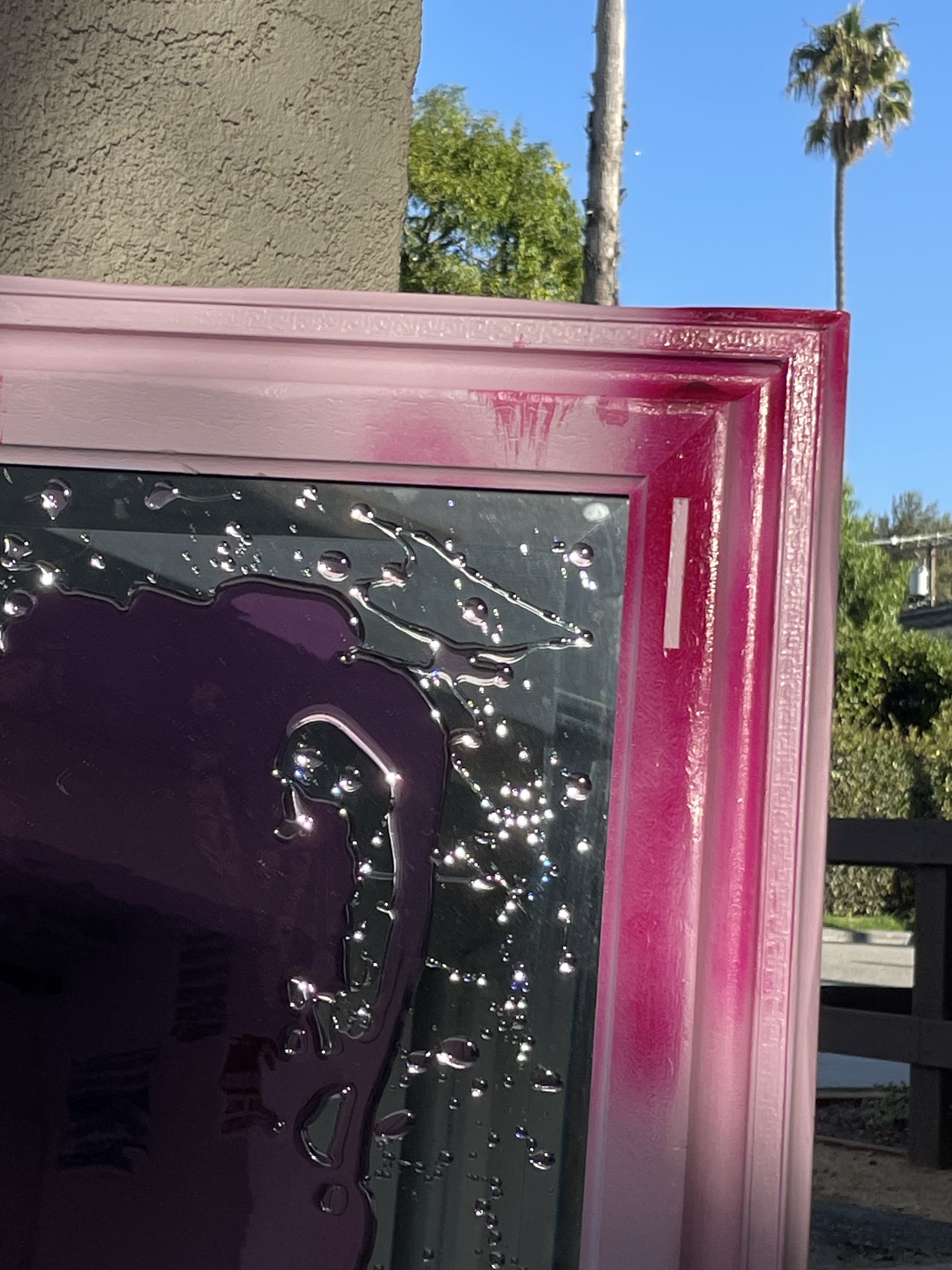 Close-up of a pink frame with some pink resin on the surface, reflecting the surroundings, with a tree, palm trees, and a blue sky in the background.